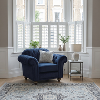 The Chesterfield Velvet Armchair in Royal Blue sits by a bay window with plants and a side table with lamp, adding timeless elegance to a bright living room. Handcrafted in England for superior quality.