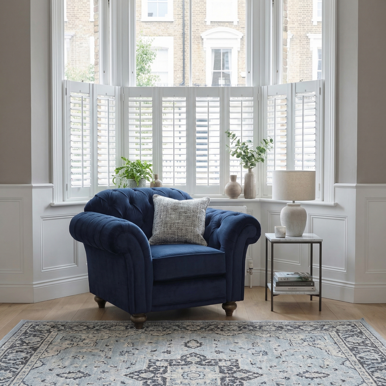The Chesterfield Velvet Armchair in Royal Blue sits by a bay window with plants and a side table with lamp, adding timeless elegance to a bright living room. Handcrafted in England for superior quality.
