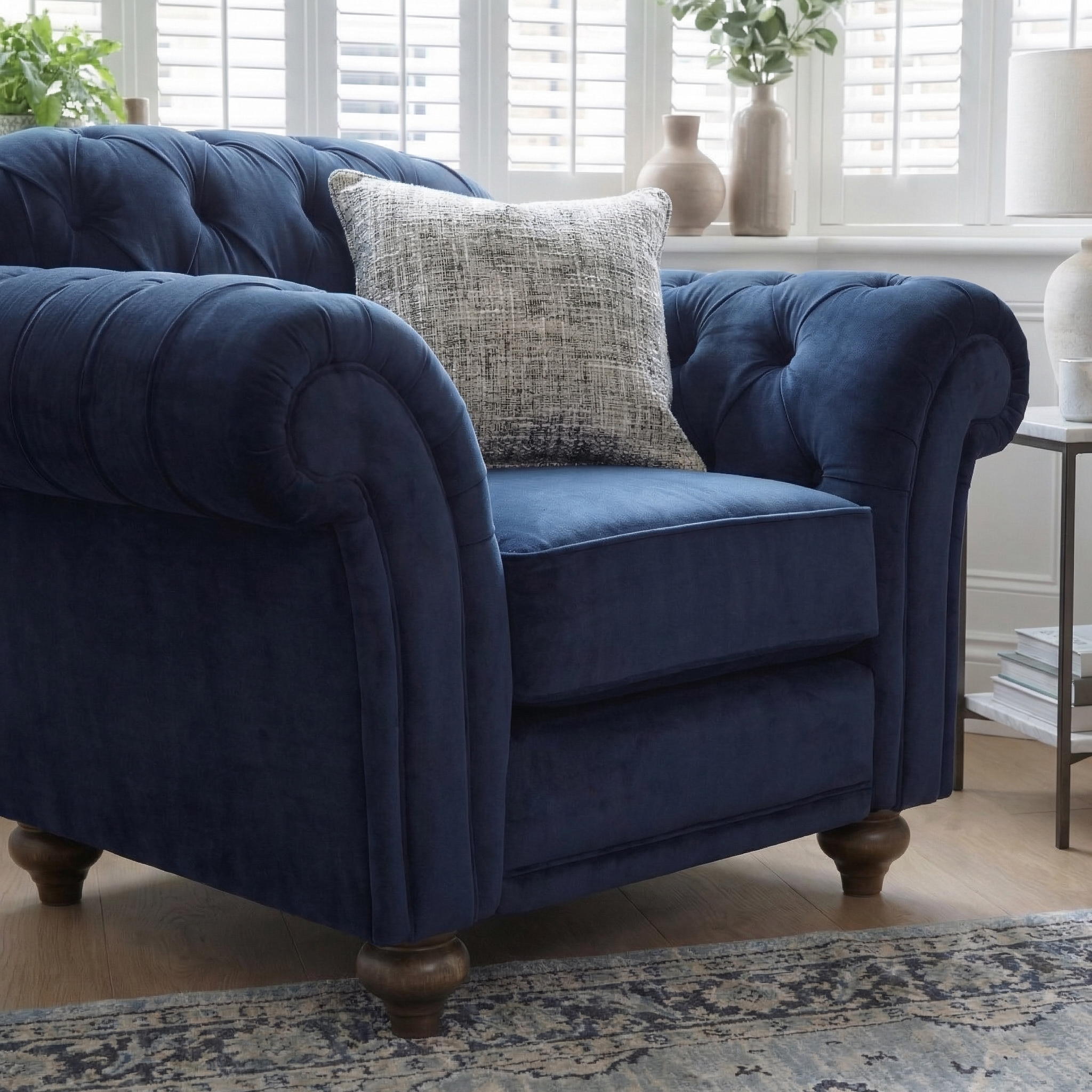 The Chesterfield Velvet Armchair in Royal Blue, handcrafted in England, features a gray pillow and sits in a bright room with white shutters and a rug.