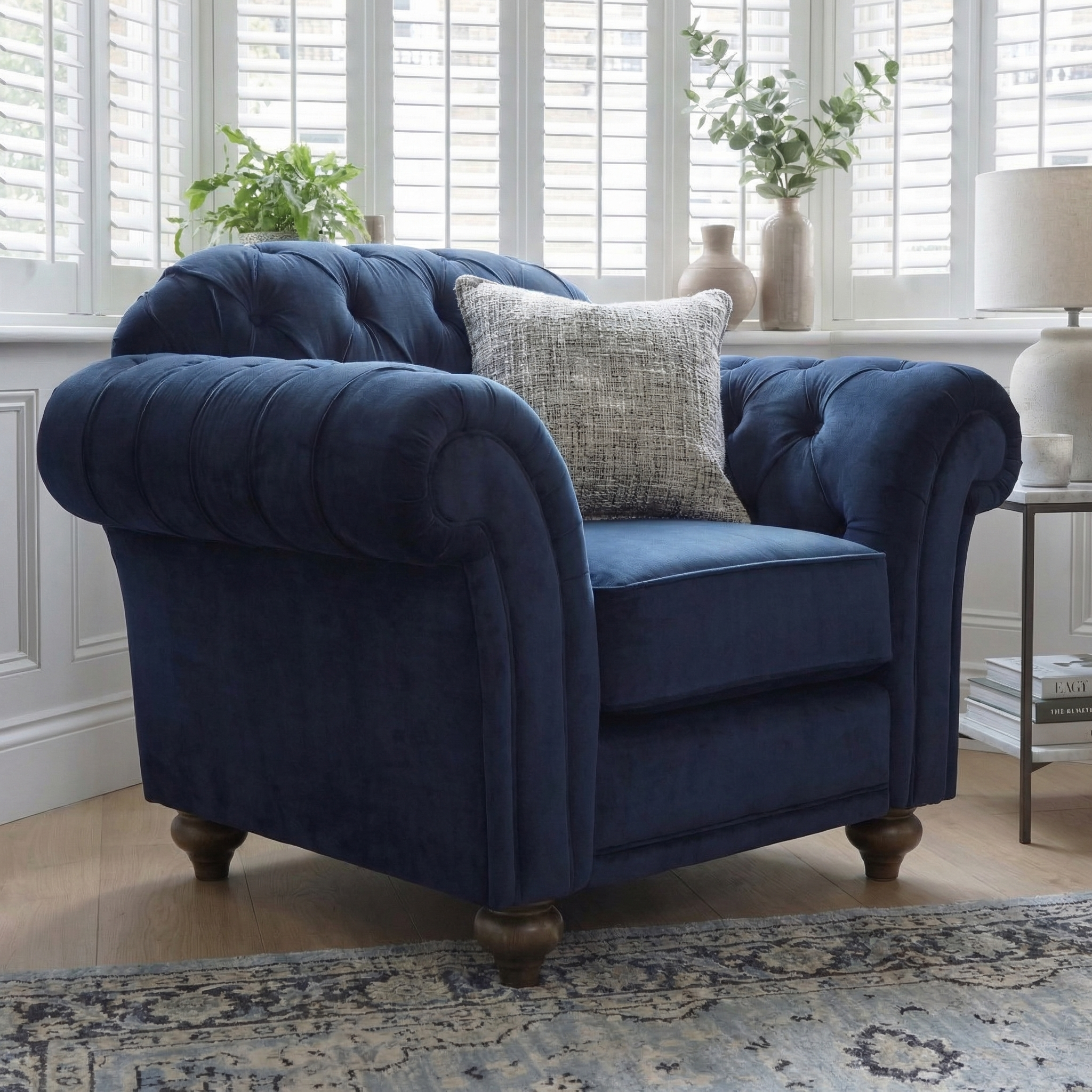 The Chesterfield Velvet Armchair in Royal Blue is handcrafted in England and features a gray cushion. Shown in a bright room with plants, a lamp, and a patterned rug.