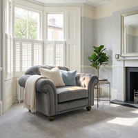 The Chesterfield Velvet Armchair in Steel Grey, accented with pillows and a blanket, brings comfort and style to a bright, elegant living room with large windows and greenery.