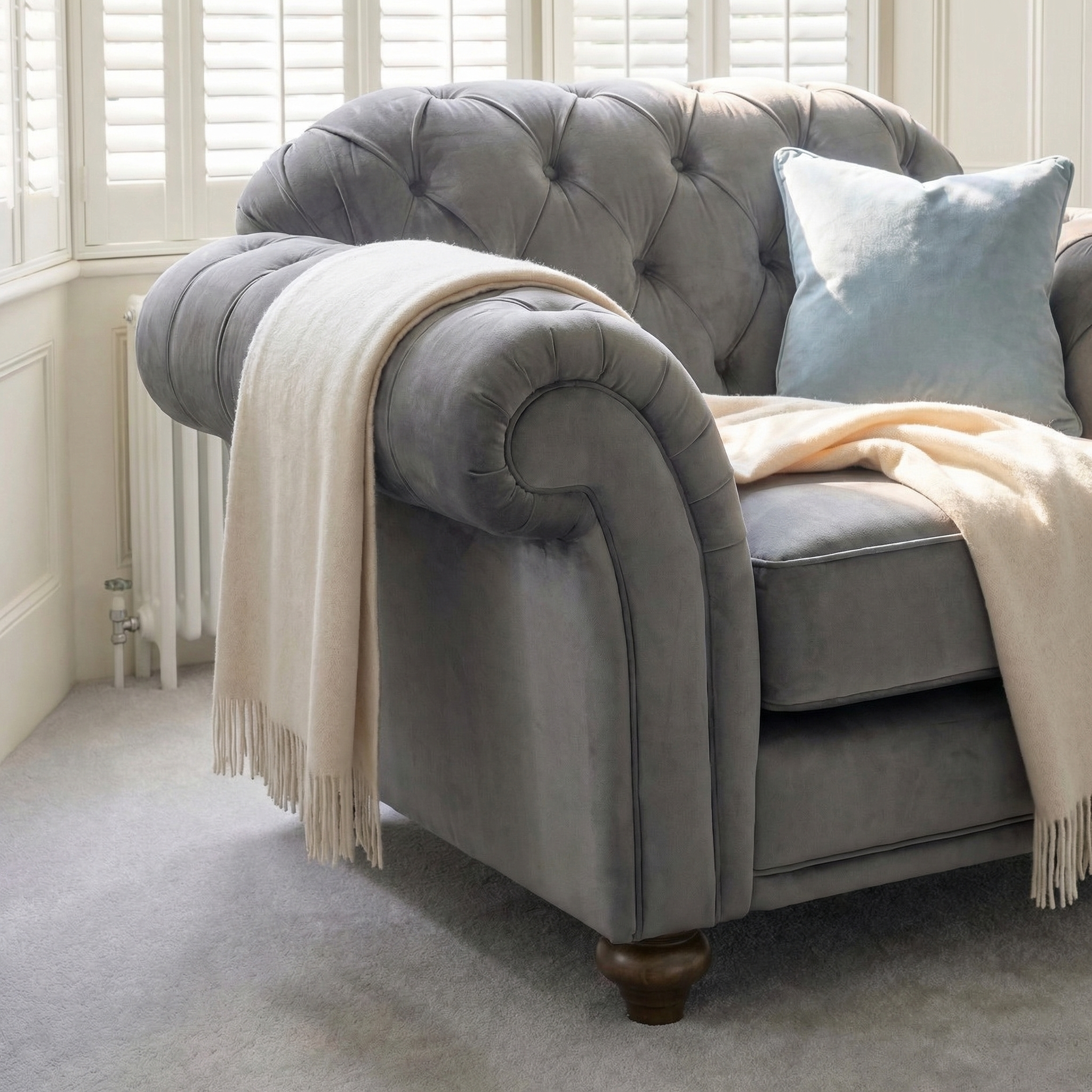 Chesterfield Velvet Armchair in Steel Grey with a light blue pillow and cream throw blanket, placed in a sunlit room with shutters.