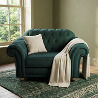 The Chesterfield Velvet Loveseat in Bottle Green, paired with a cream pillow and throw blanket by the window on a green patterned rug, brings refined elegance reminiscent of a classic handcrafted sofa.