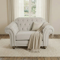 The Chesterfield Velvet Loveseat in Feather Grey sits on a rug before a window with sheer curtains, accented by a beige throw and pillow.