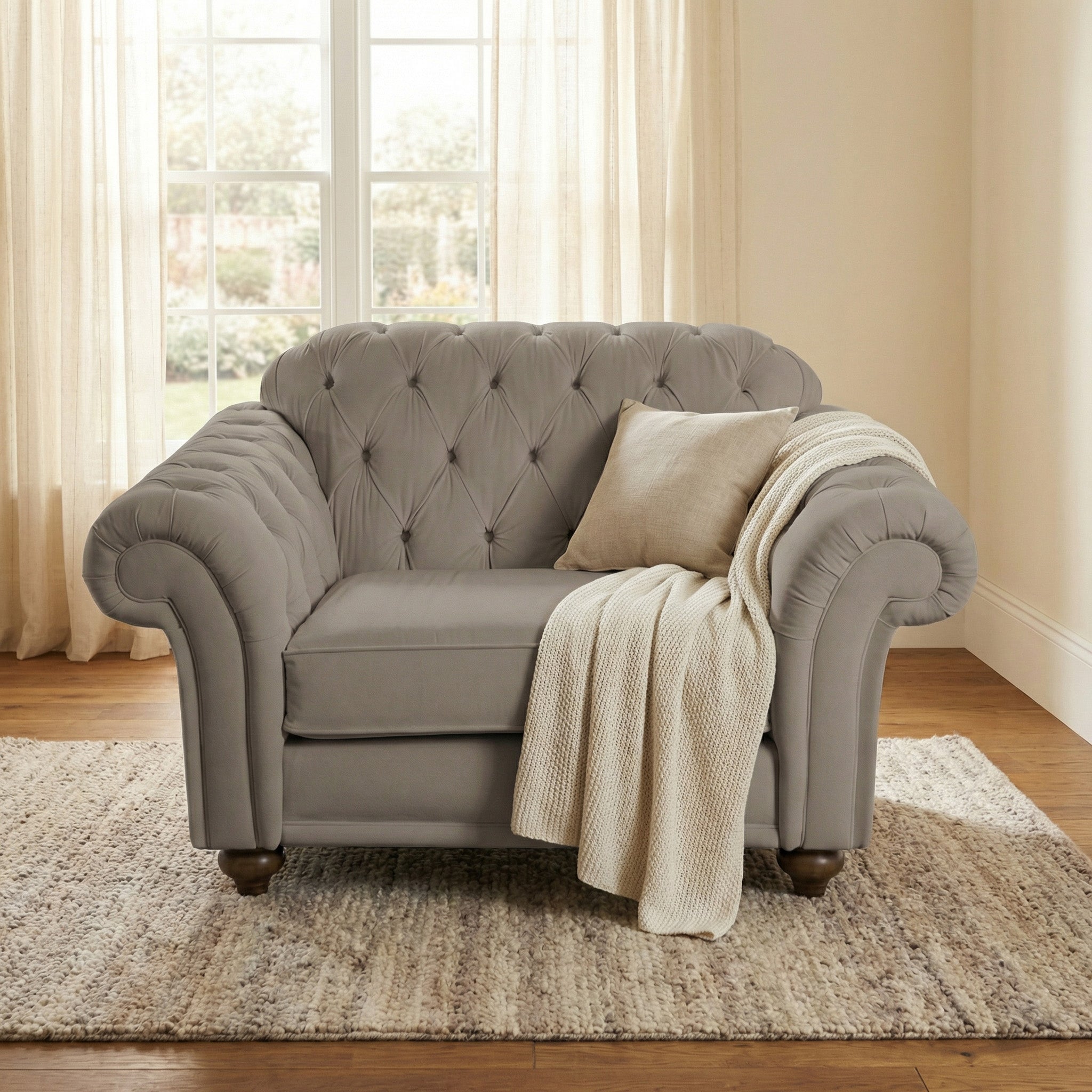Gray tufted armchair with a beige pillow and knit blanket on a rug, styled beside a Chesterfield Velvet Loveseat in Mink, set in front of tall windows with sheer curtains.