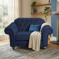 The Chesterfield Velvet Loveseat in Royal Blue, adorned with a blue pillow and cream knit throw, rests on a rug in a cozy, sunlit room.