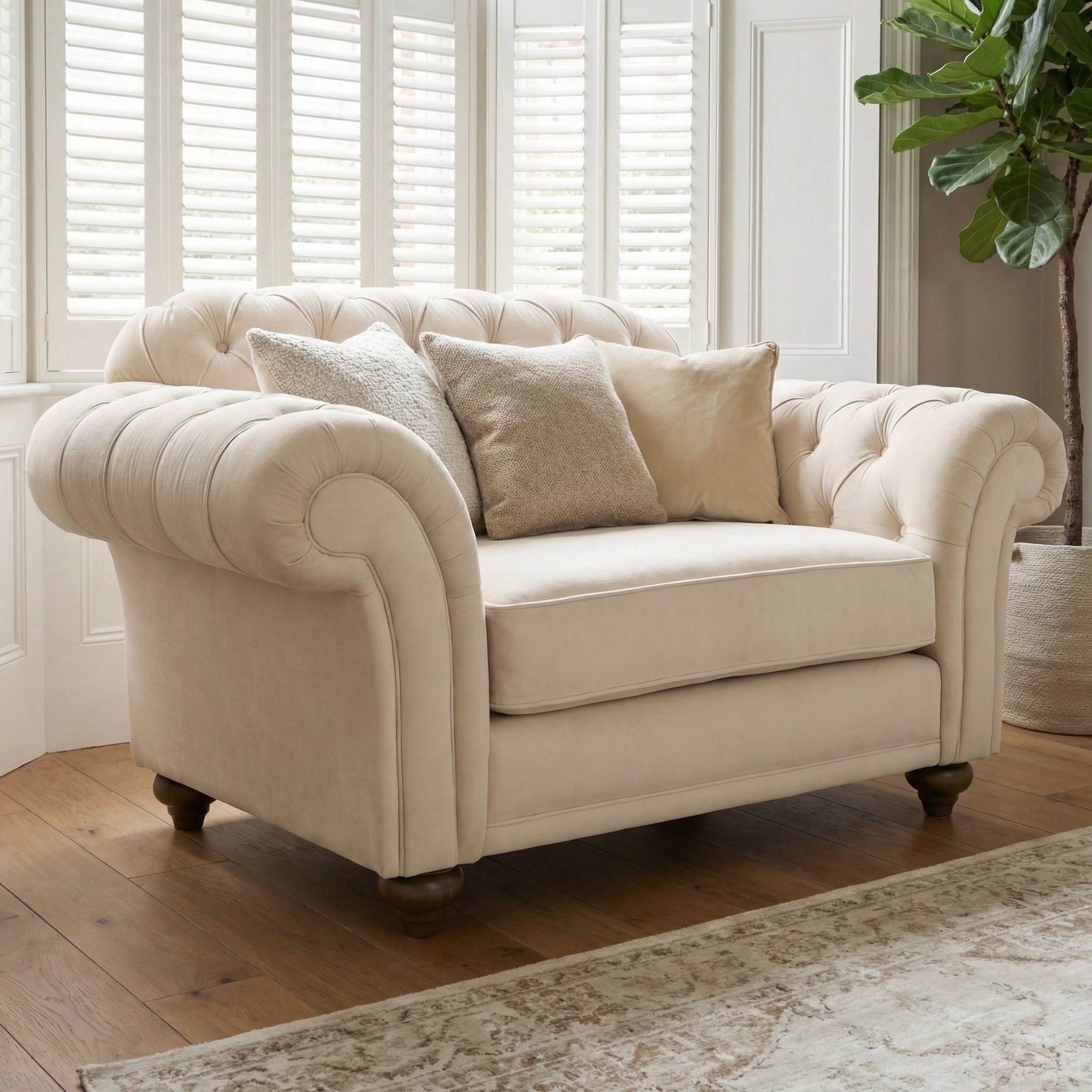 The Chesterfield Velvet Loveseat in Cream features plush, tufted cushions and is shown on a wooden floor by windows with white shutters.