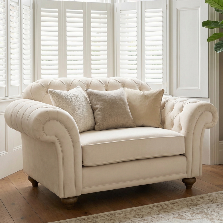 The Chesterfield Velvet Loveseat in Cream, handcrafted in England, sits in a bright room with wooden floors, shuttered windows, and three neutral pillows.