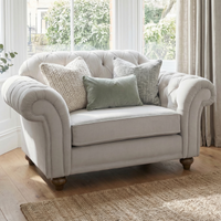 A Chesterfield Velvet Loveseat in Feather Grey, accented with four decorative pillows, sits by large windows in a bright, cozy room.