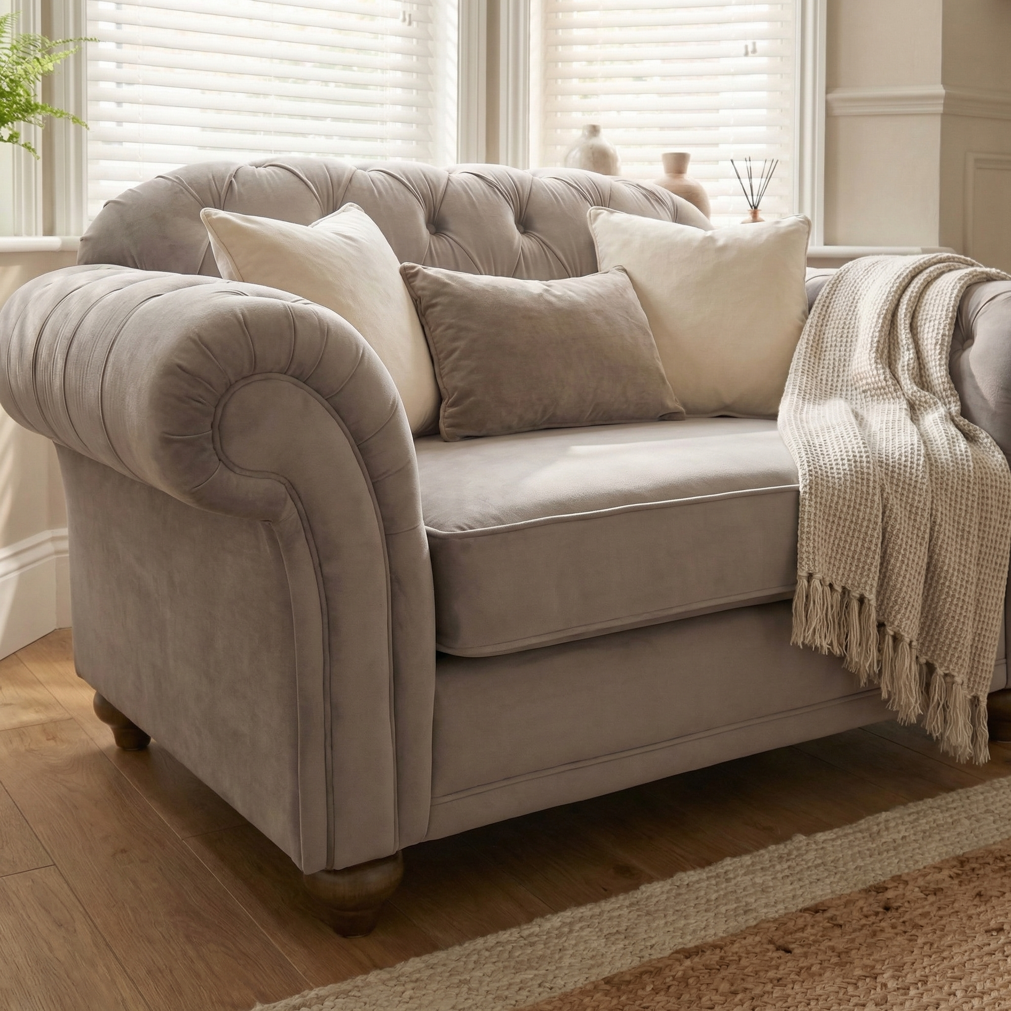 Chesterfield Velvet Loveseat in Mink featuring cream pillows and a knit throw, placed in a bright room with wooden floors and window blinds.