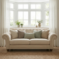 The Chesterfield 2 Seat Velvet Sofa in Cream, adorned with assorted neutral pillows, is placed in front of large bay windows dressed in white curtains, while plants decorate the windowsill.