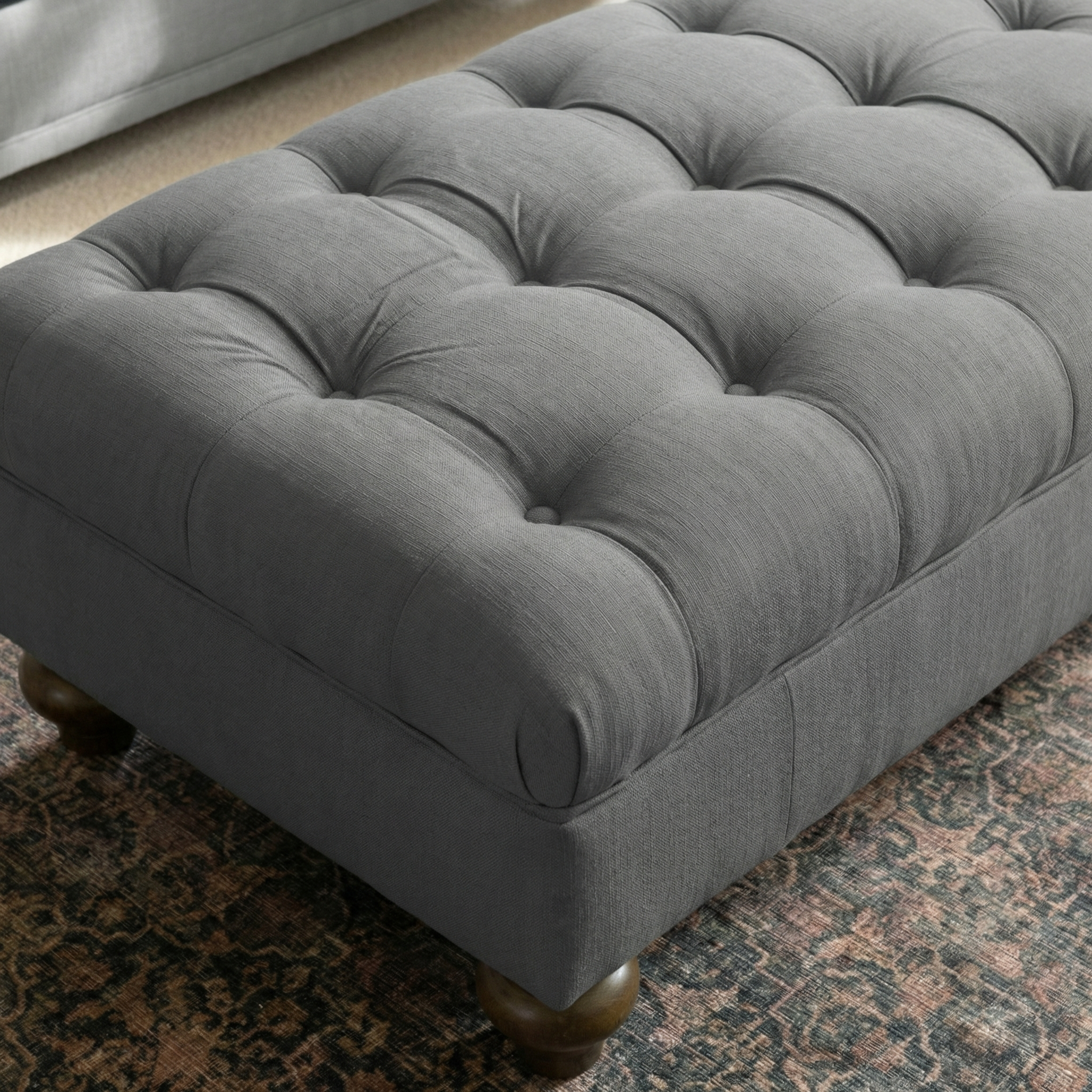 The Chesterfield Footstool in Smoke Grey with wooden legs sits on a patterned rug, showcasing UK craftsmanship; a sofa is partially visible in the background.