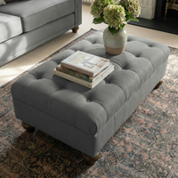 The Chesterfield Footstool in Smoke Grey, handcrafted in the UK, is topped with books and a vase of green hydrangeas and sits on a patterned rug.