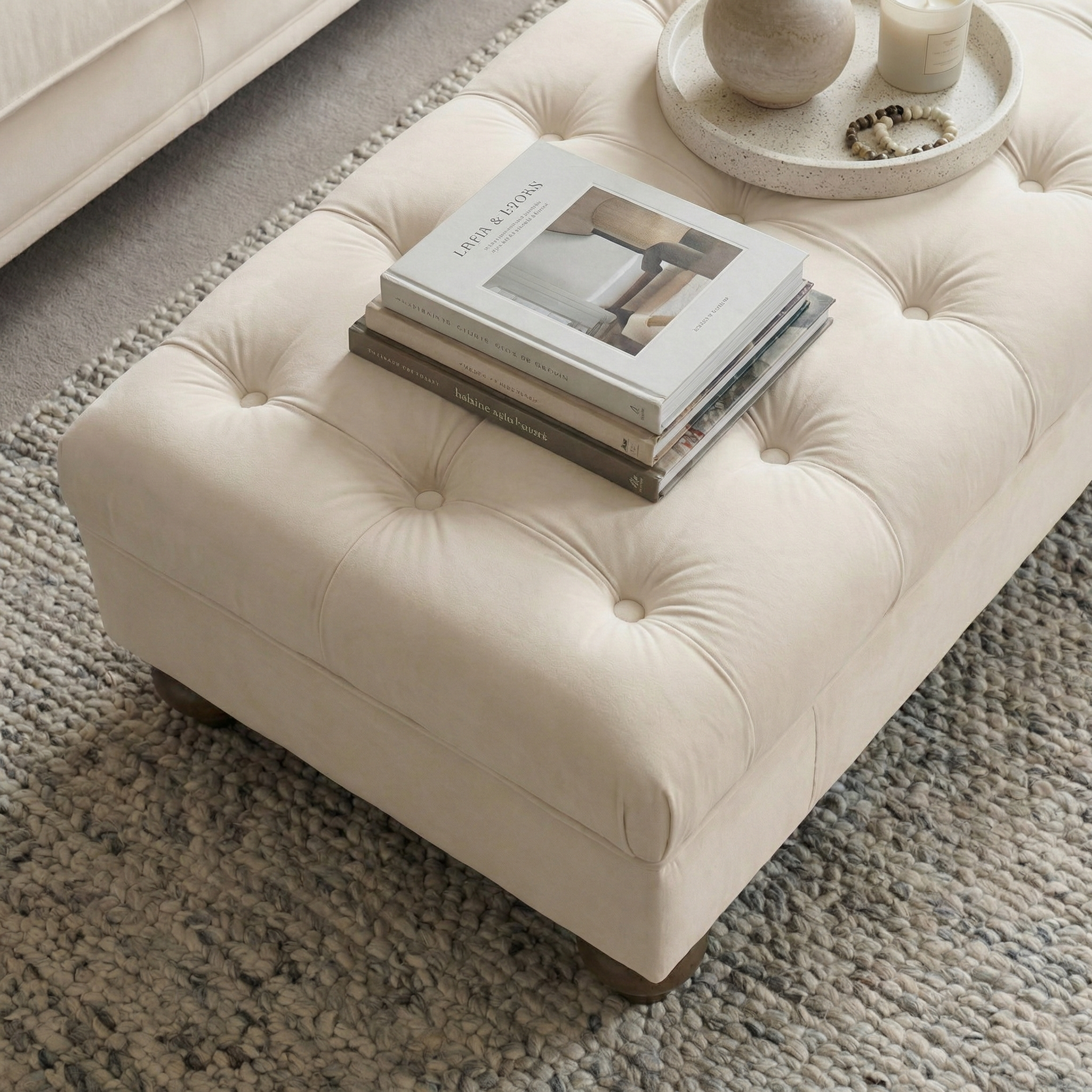 The Chesterfield Velvet Footstool in Cream displays stacked books and a tray with candles and decor, all resting on a textured rug.