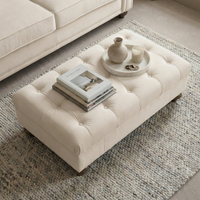 The Chesterfield Velvet Footstool in Cream holds books, candles, and vases on a tray, set atop a textured rug in a cozy living room.