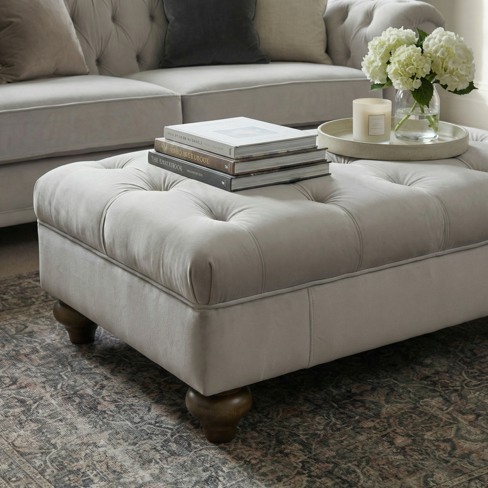 The Chesterfield Velvet Footstool in Feather Grey serves as a coffee table in the living room, showcasing stacked books, a candle, and flowers arranged on a tray.