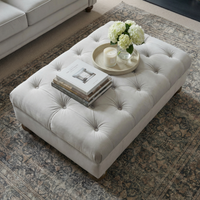 The Chesterfield Velvet Footstool in Feather Grey, styled with books, a candle, and white flowers on a tray atop a patterned rug, brings handcrafted UK elegance and charm to any space.