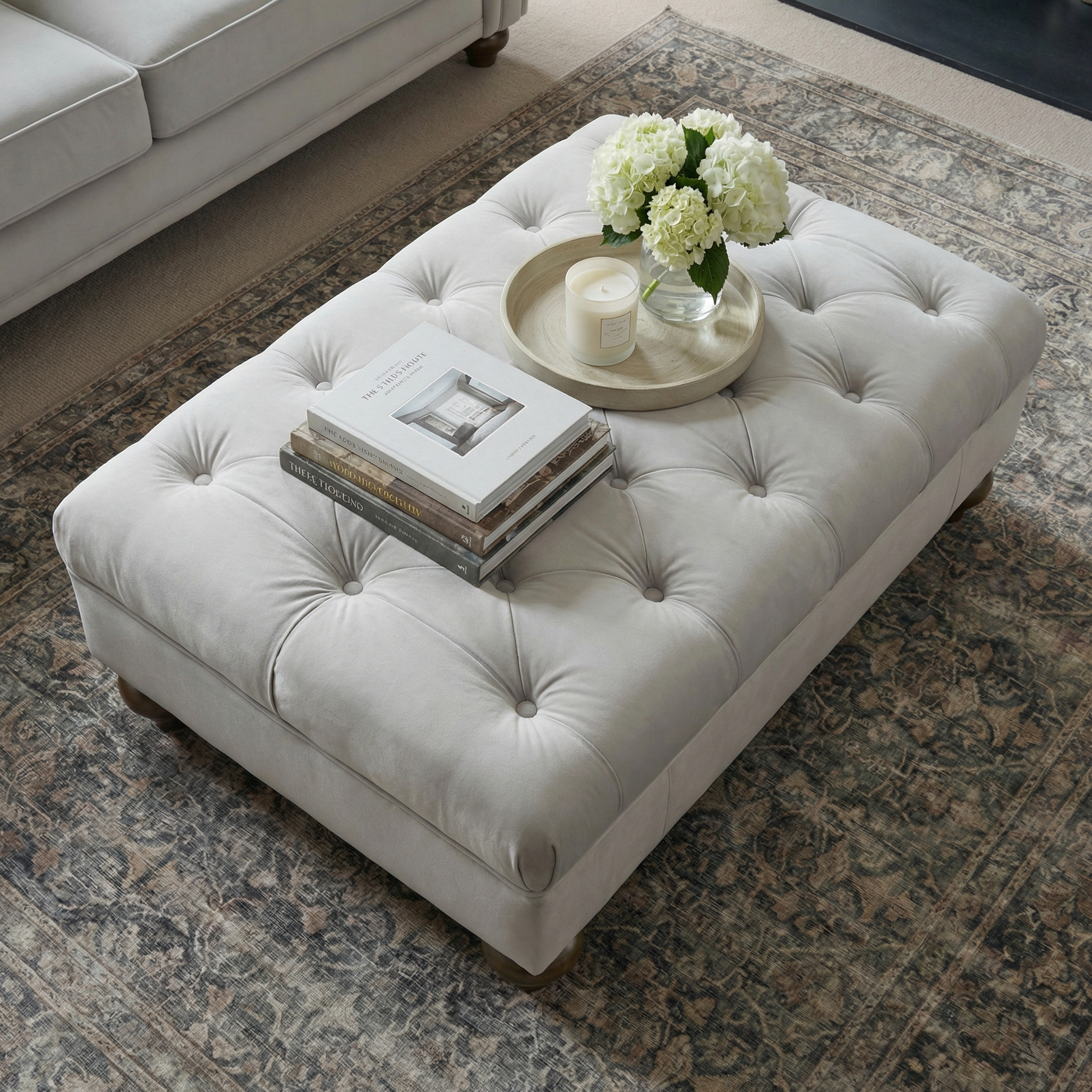 The Chesterfield Velvet Footstool in Feather Grey, styled with books, a candle, and white flowers on a tray atop a patterned rug, brings handcrafted UK elegance and charm to any space.