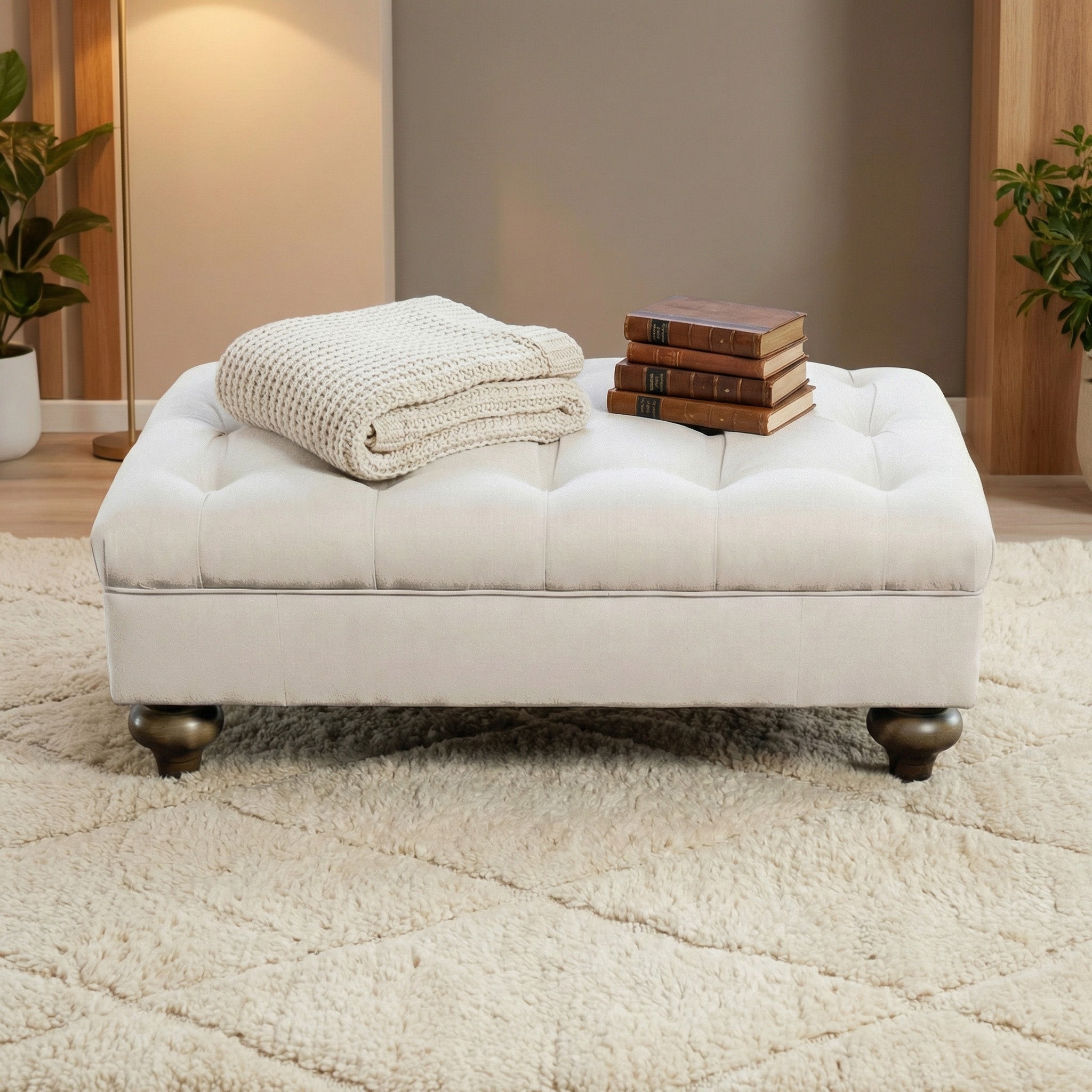 The Chesterfield Footstool in Ivory, topped with a folded knit blanket and three stacked books, rests on a plush beige rug.
