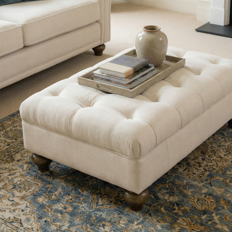 The Chesterfield Footstool in Ivory, topped with a tray of books and a vase, sits on a patterned rug in the living room.