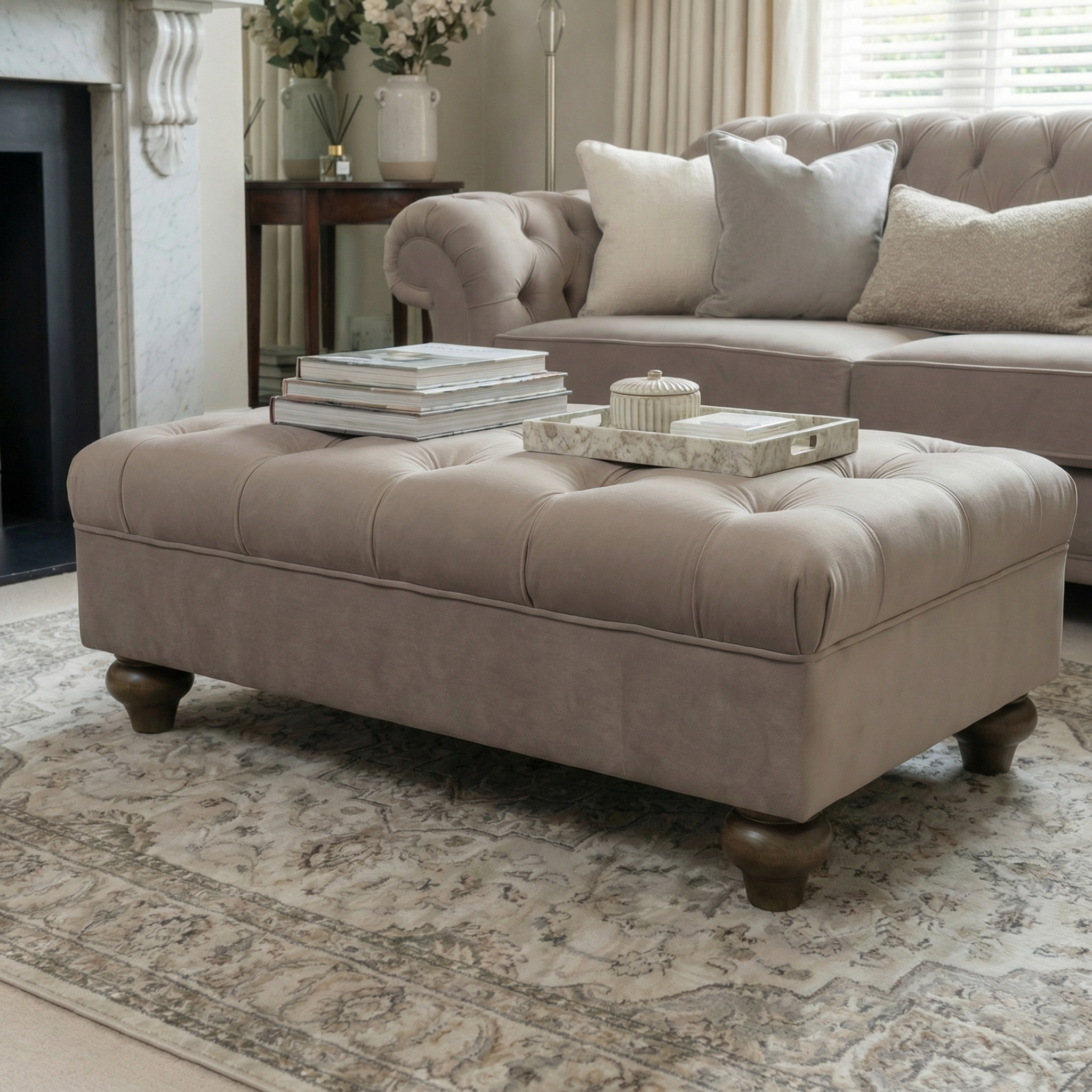 The Chesterfield Velvet Footstool in Mink, topped with books and a tray, sits on a patterned rug in a cozy living room.