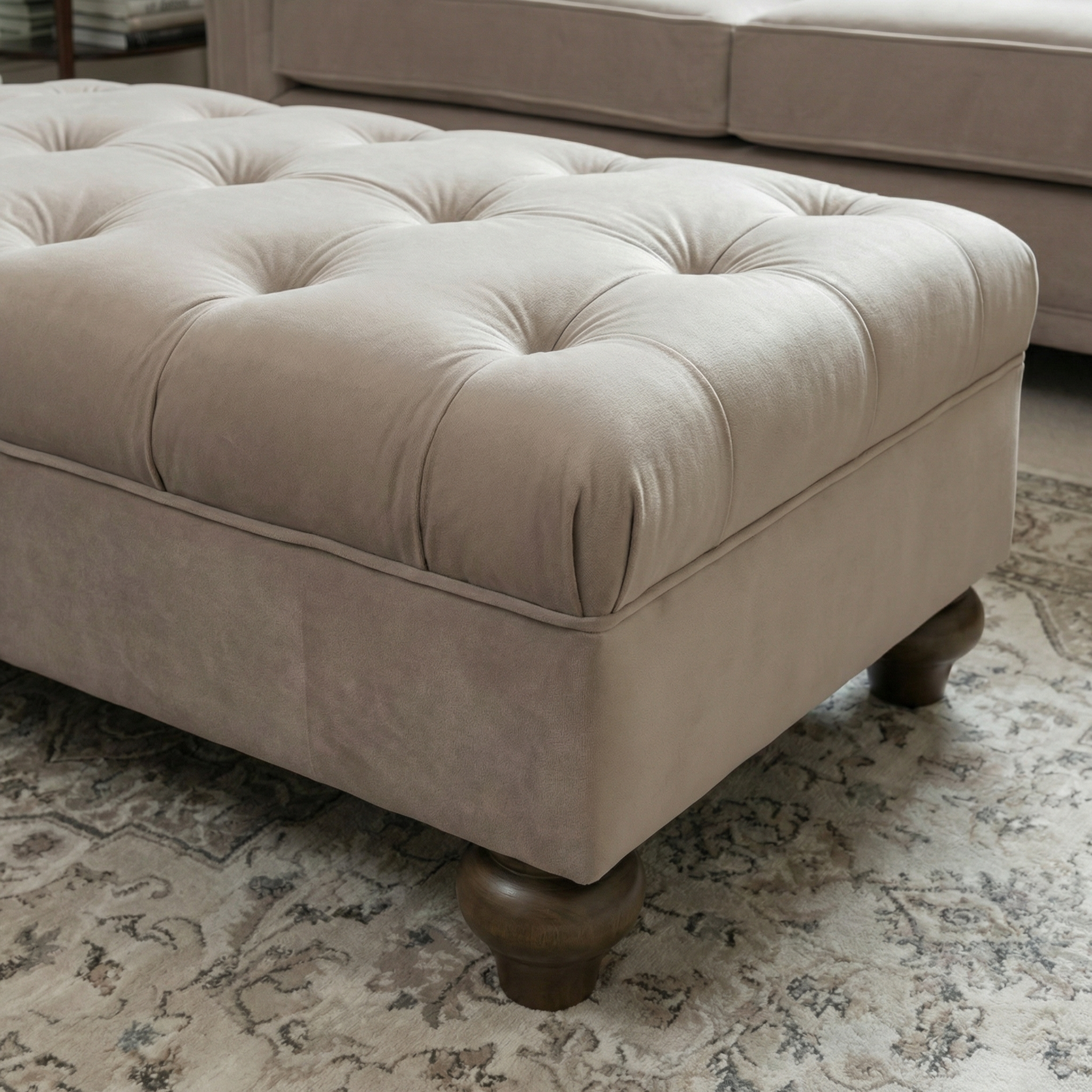 The Chesterfield Velvet Footstool in Mink, with elegant wooden legs, sits on a patterned rug beside a matching beige sofa, adding a refined touch to the room.