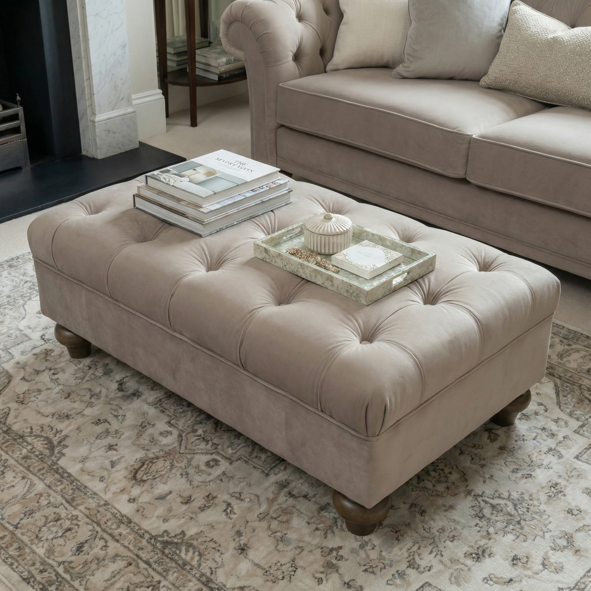 The Chesterfield Velvet Footstool in Mink, topped with books and a decorative tray, sits on a patterned rug in a cozy living room.