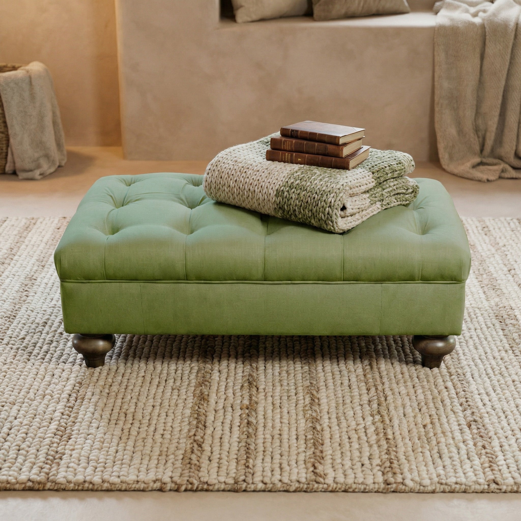 The Chesterfield Footstool in Olive, topped with knitted blankets and books, sits on a textured rug in a cozy room, showcasing classic British design charm.