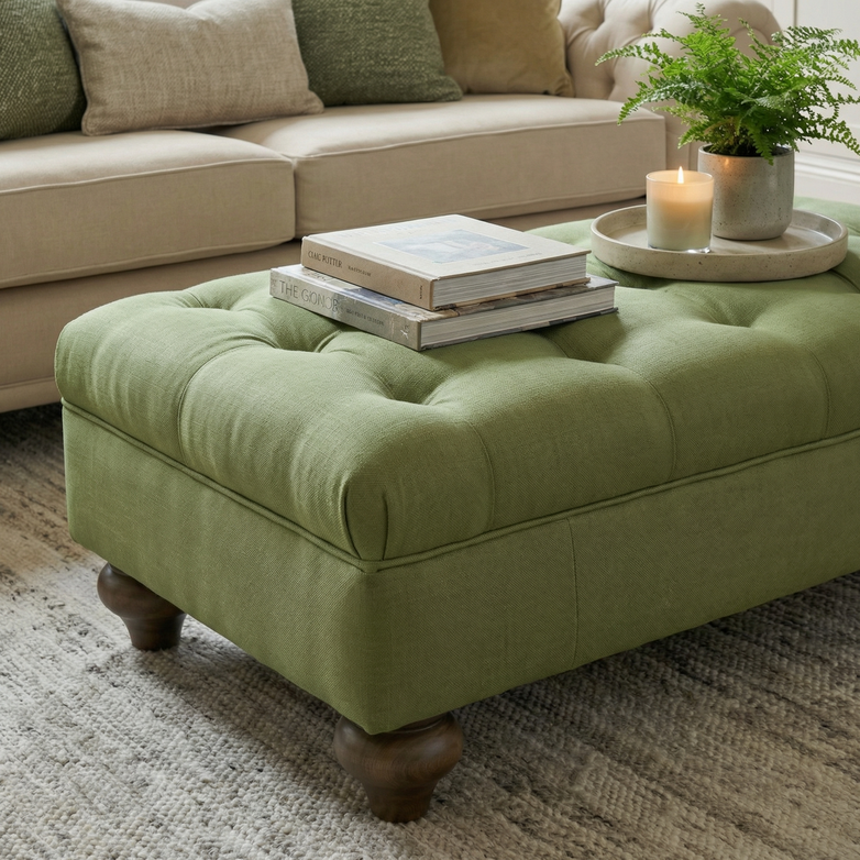 The Chesterfield Footstool in Olive, adorned with books, a lit candle, and a potted fern, sits before a beige sofa—an inviting nod to British design.