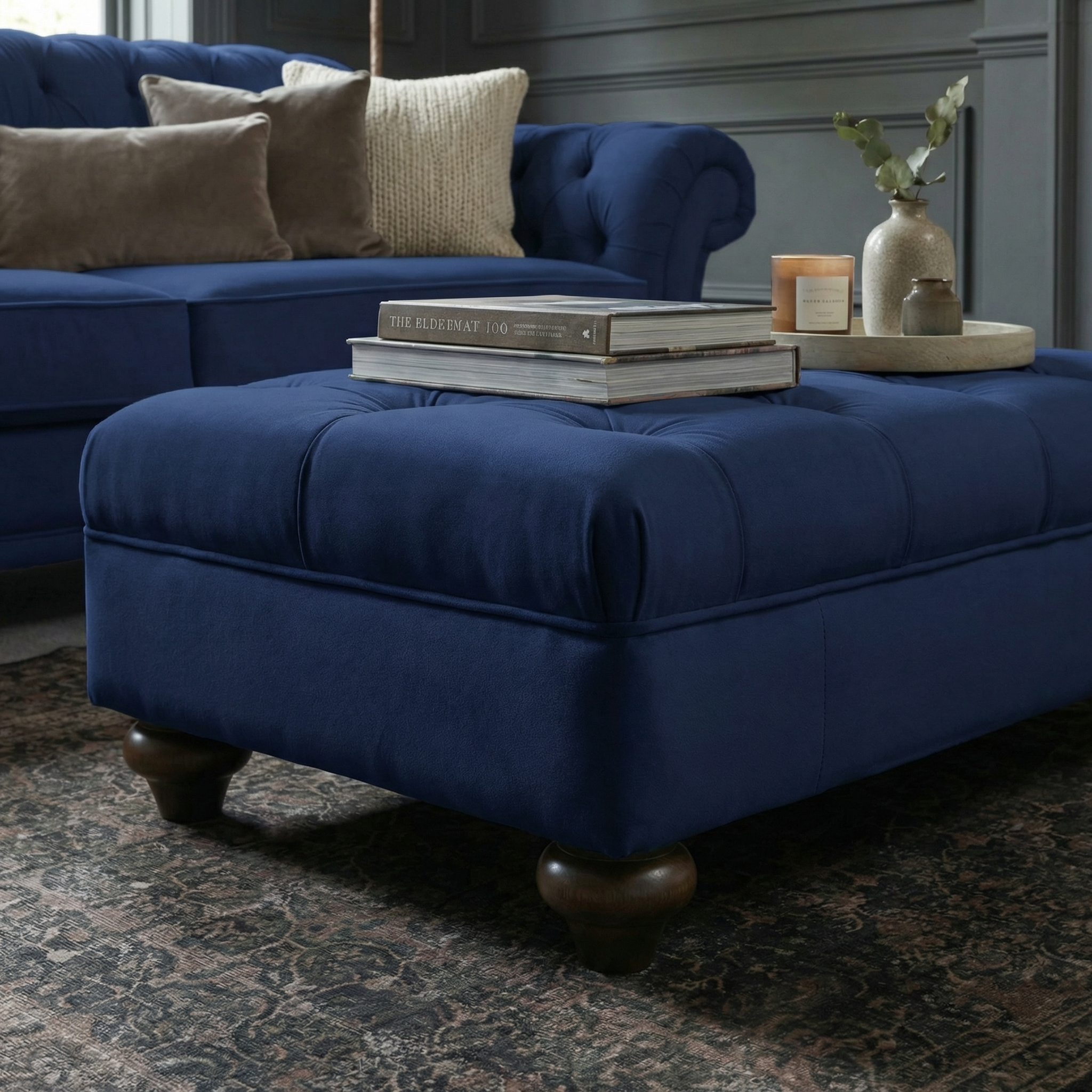 The Chesterfield Velvet Footstool in Royal Blue, topped with books and decor, sits before a matching sofa and adds charm to this cozy living room.