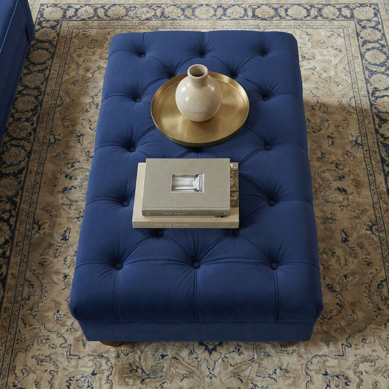 The Chesterfield Velvet Footstool in Royal Blue is styled with books, a candle, and a vase on a tray, set atop a patterned rug.