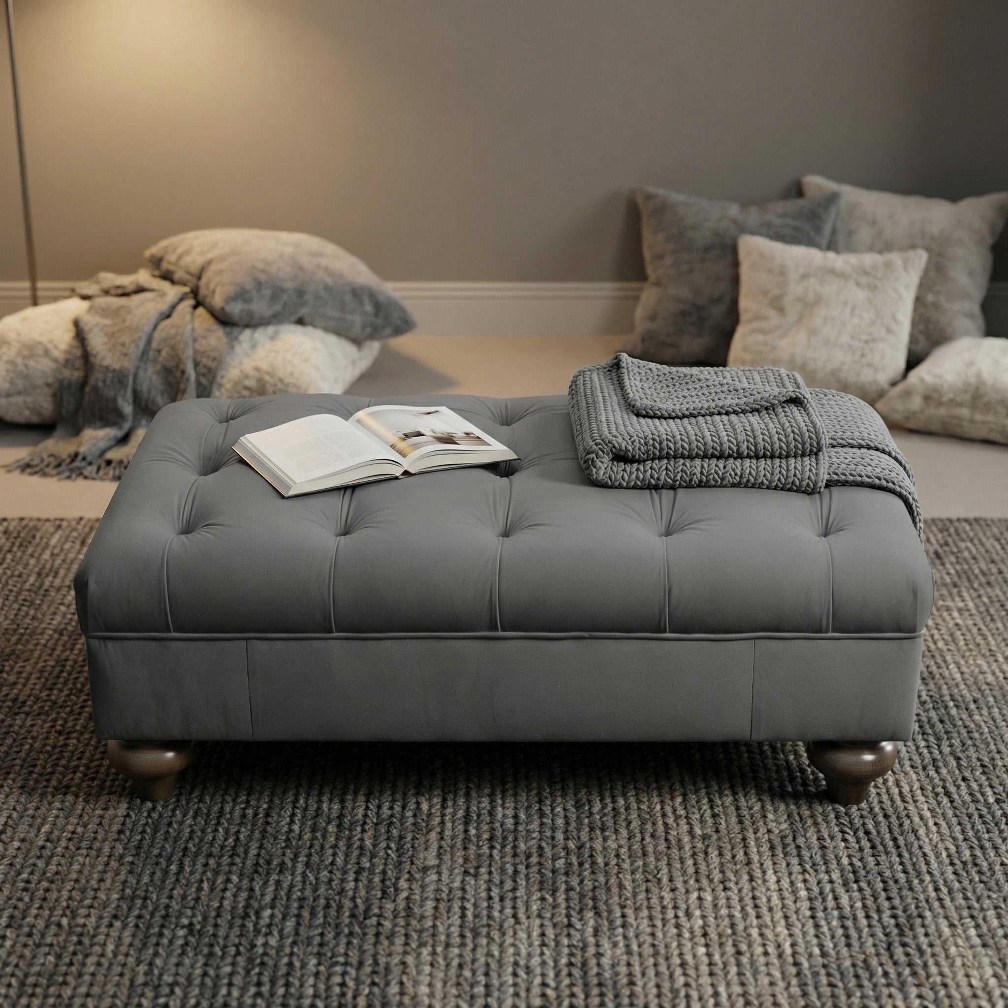 The Chesterfield Velvet Footstool in Steel Grey is shown with an open book, stacked magazines, and folded knit blankets in a cozy, softly lit room.