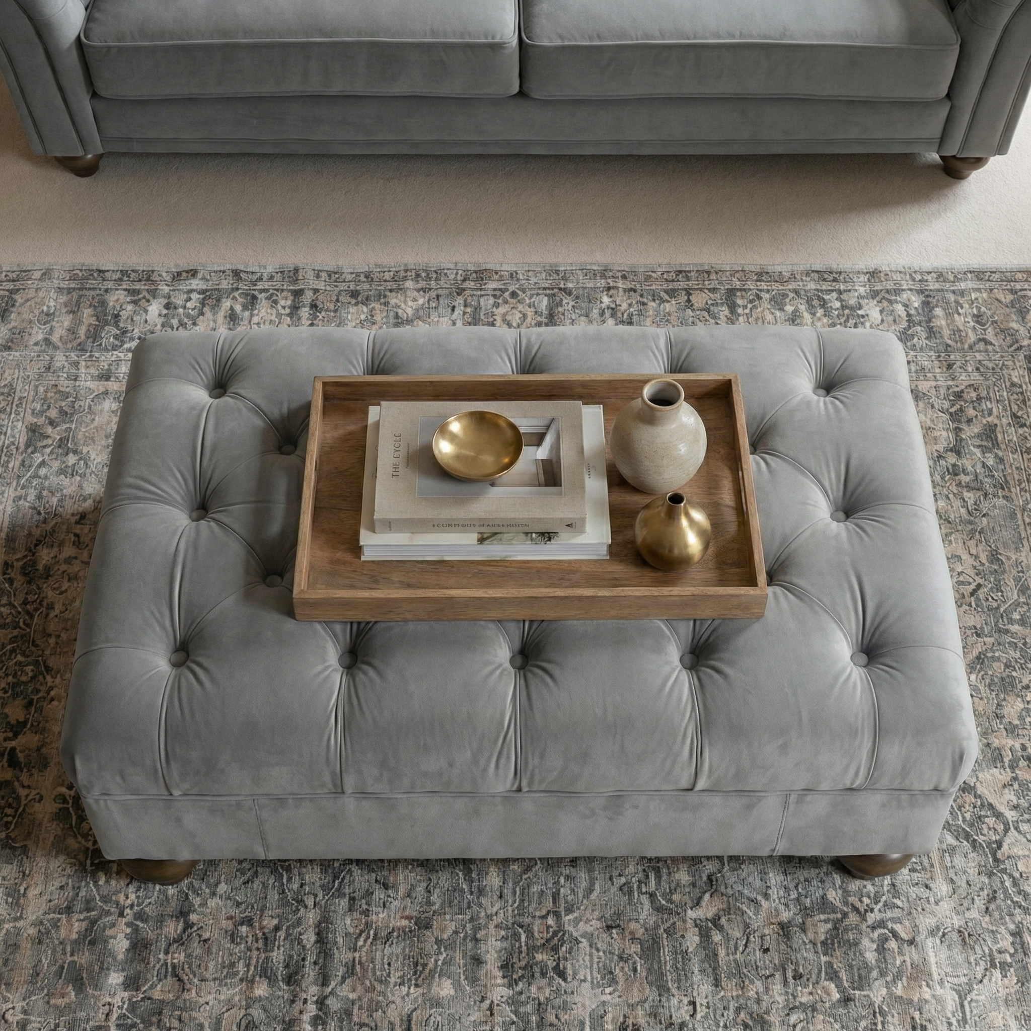 The Chesterfield Velvet Footstool in Steel Grey is topped with a wooden tray holding books, a vase, and two small gold bowls, all set on a patterned rug.