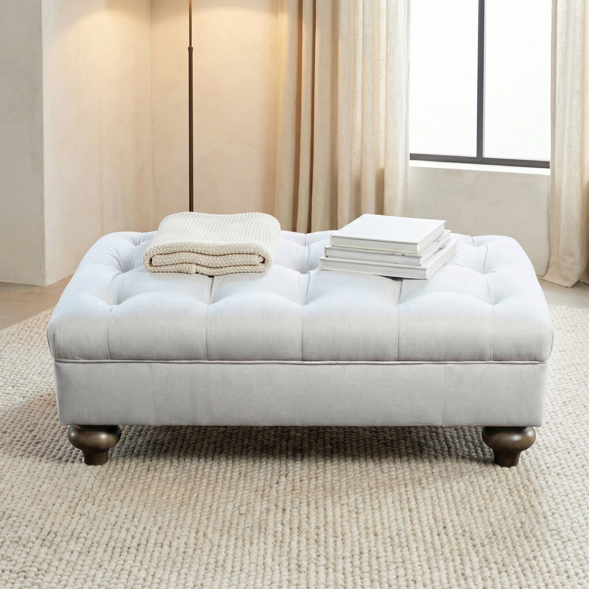 The Chesterfield Footstool in Warm Grey features traditional tufted upholstery, topped with a folded blanket and stacked books, and is set on a textured rug.