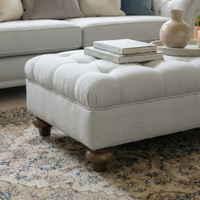 The Chesterfield Footstool in Warm Grey, topped with books and decor, is set on a patterned rug beside a sofa.