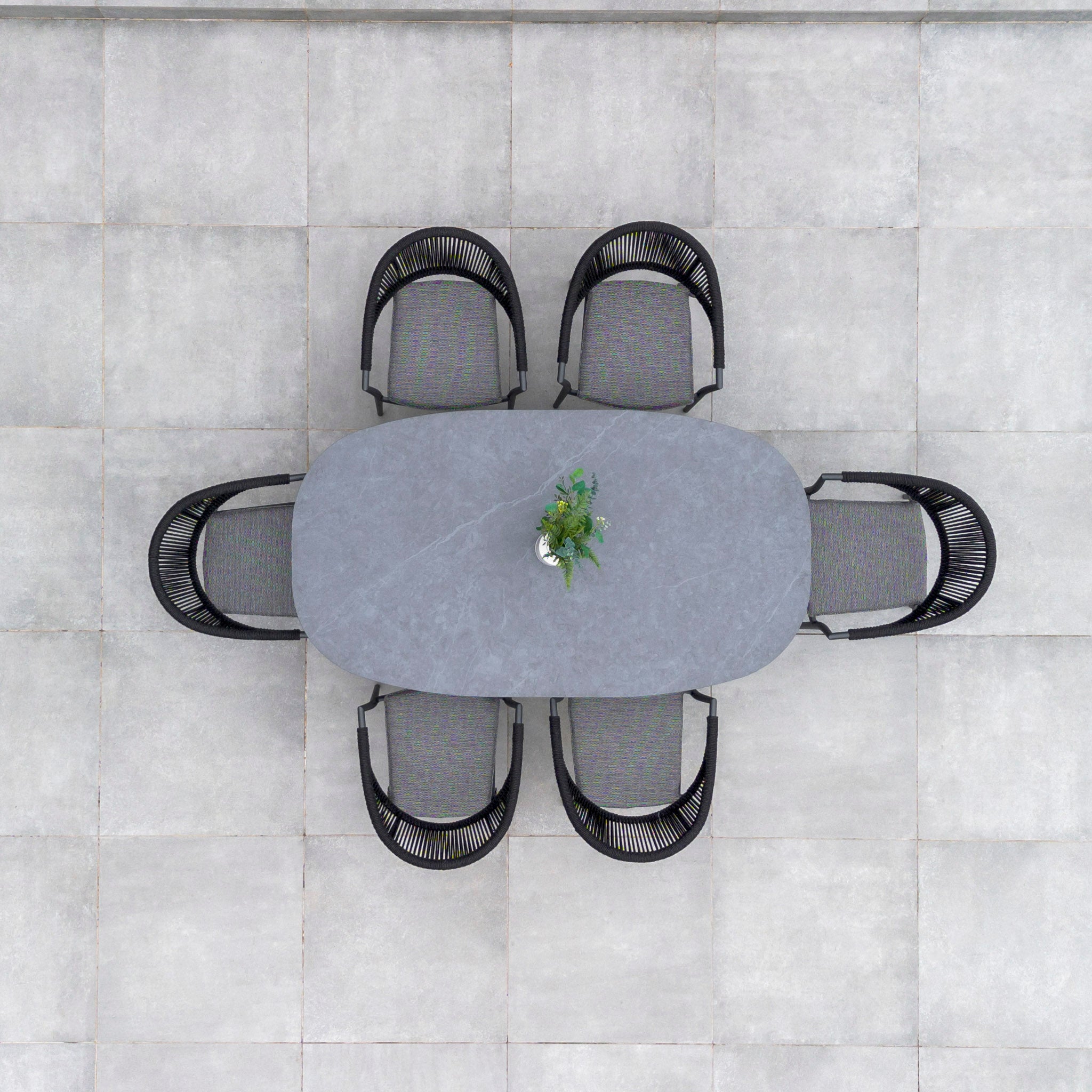 Top view of the Cloverly 6 Seat Rope Oval Dining Set with Ceramic Table in Charcoal, featuring six woven chairs and a potted plant centerpiece on a tiled patio.