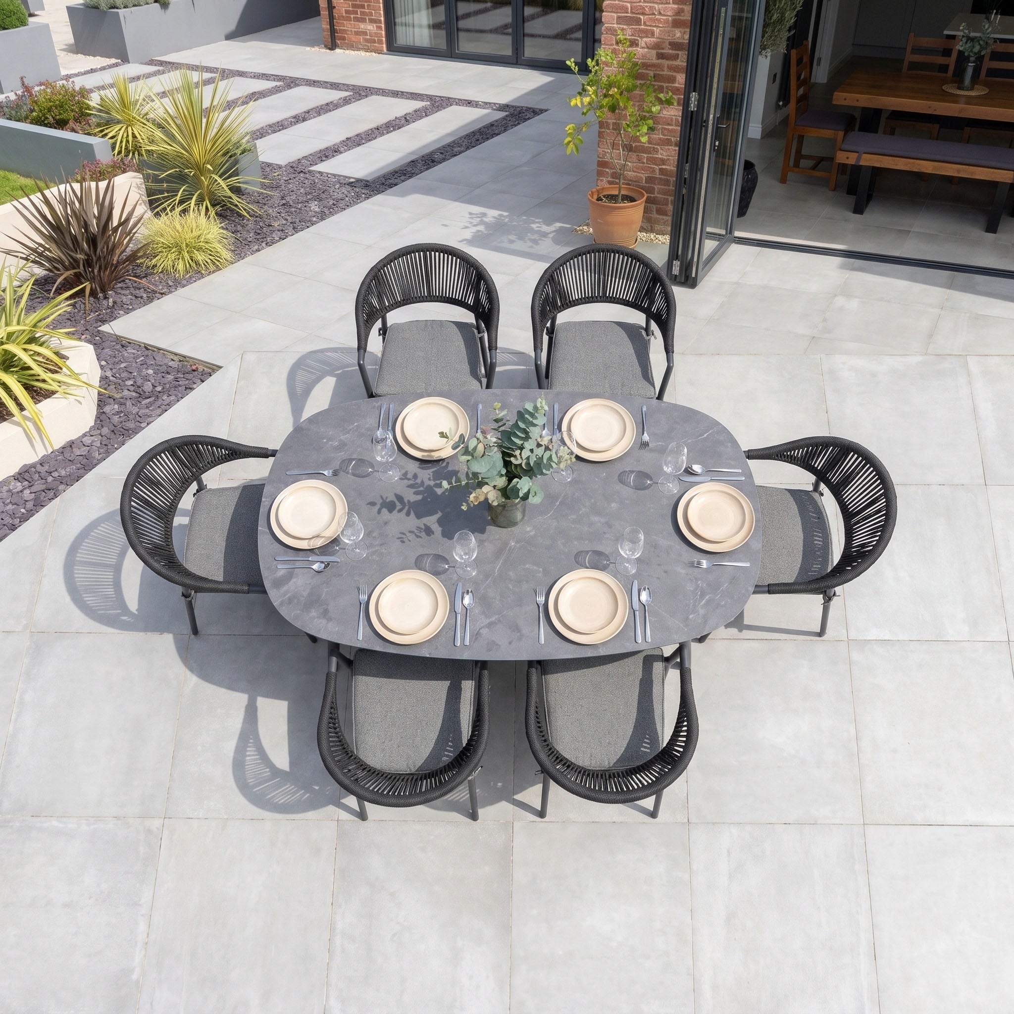 The Cloverly 6 Seat Oval Dining Set with Albatross Table in Charcoal creates an inviting patio setup for six, featuring stackable dining chairs for convenient storage and a stylish arrangement with neutral plates, cutlery, and a plant centerpiece.