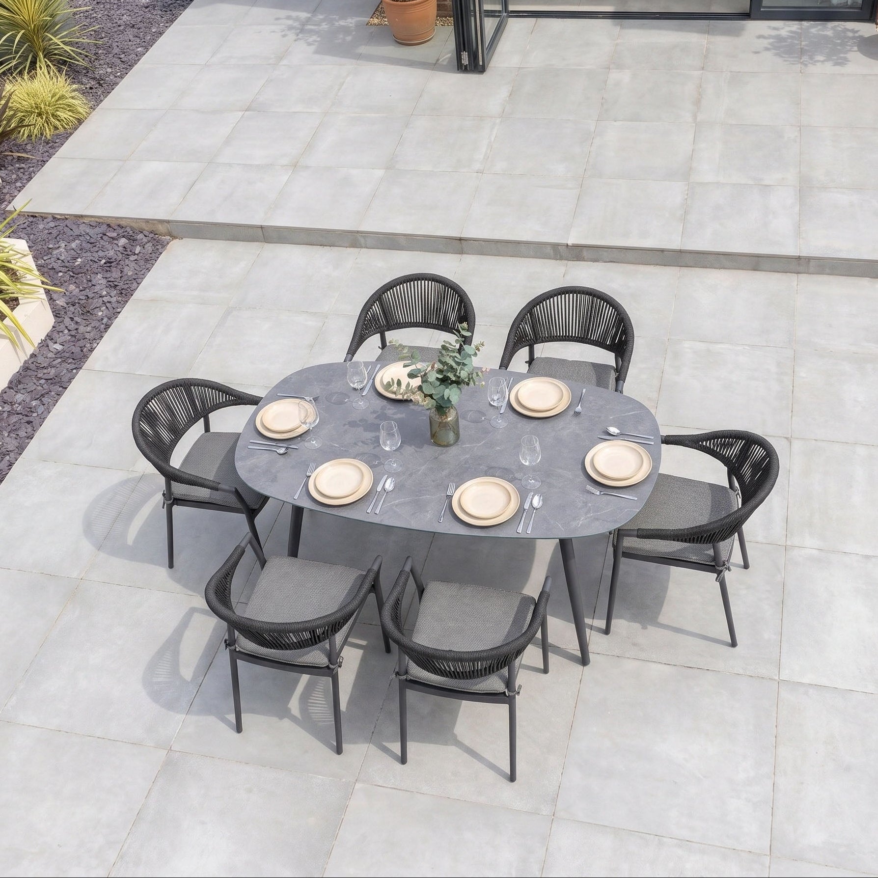 The Cloverly 6 Seat Oval Dining Set with Albatross Table in Charcoal features plates and glasses arranged on its spacious tabletop, surrounded by stylish black stackable dining chairs on a tiled patio. This elegant set creates the perfect setting for memorable outdoor gatherings.