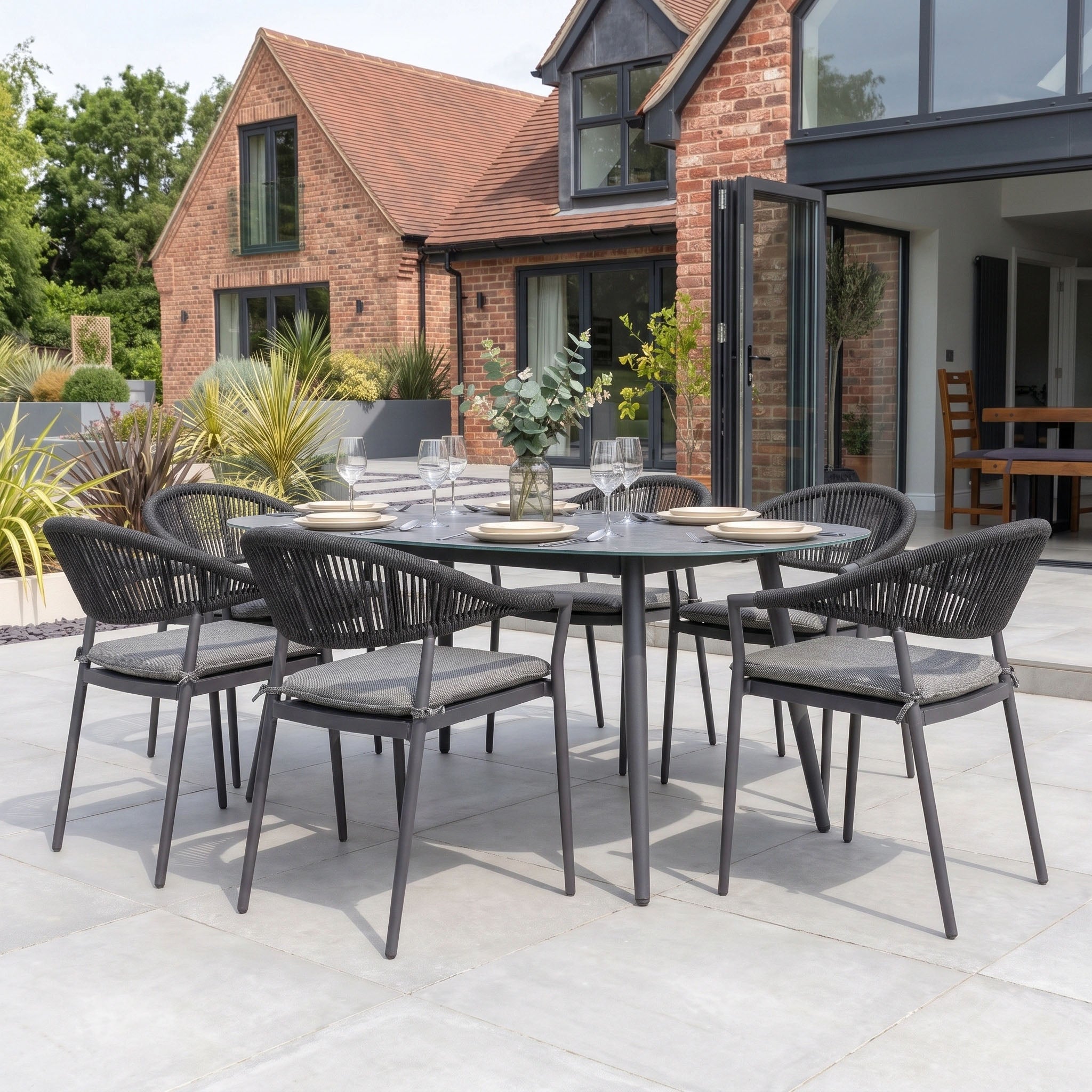 The Cloverly 6 Seat Oval Dining Set with Albatross Table in Charcoal, featuring stackable dining chairs, is displayed on a patio in front of a contemporary brick house.