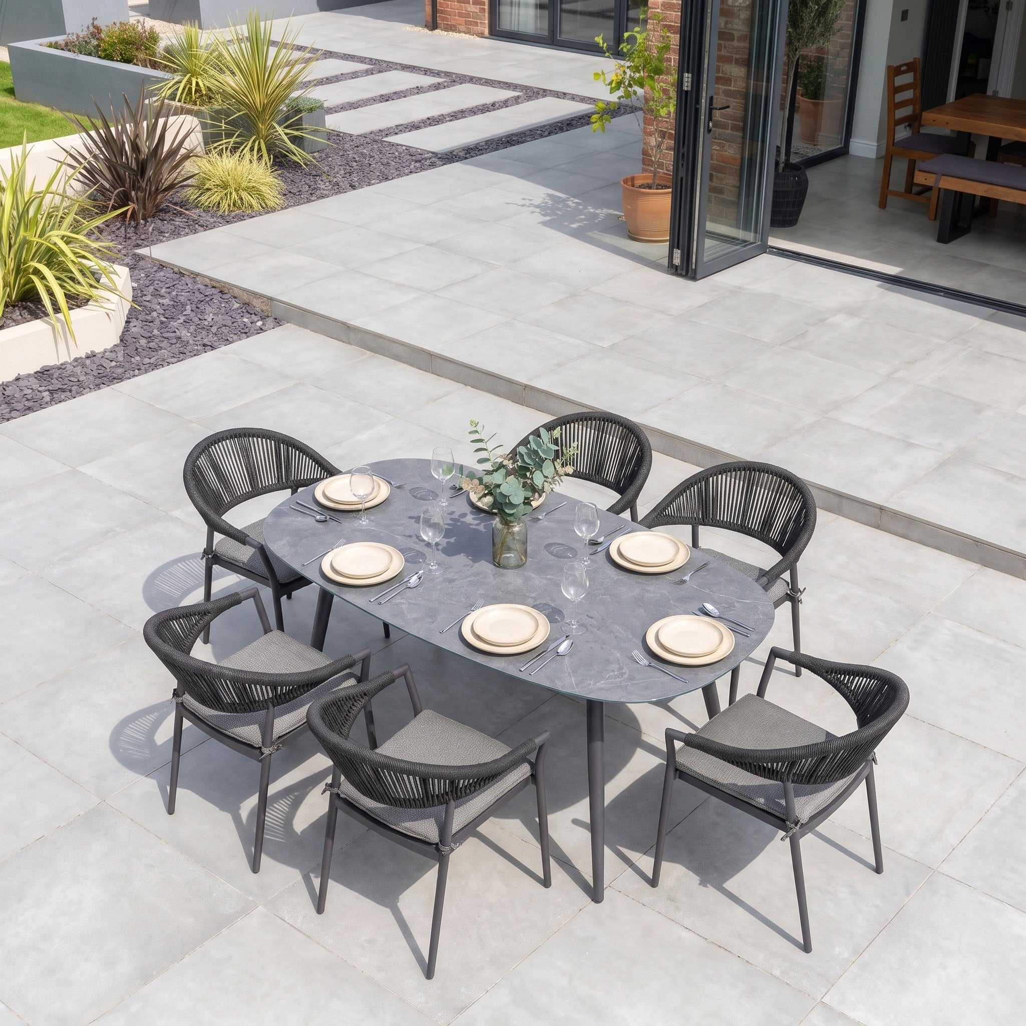 Cloverly 6 Seat Oval Dining Set with Albatross Table in Charcoal, featuring stackable dining chairs and arranged with plates and a vase on a tiled patio.