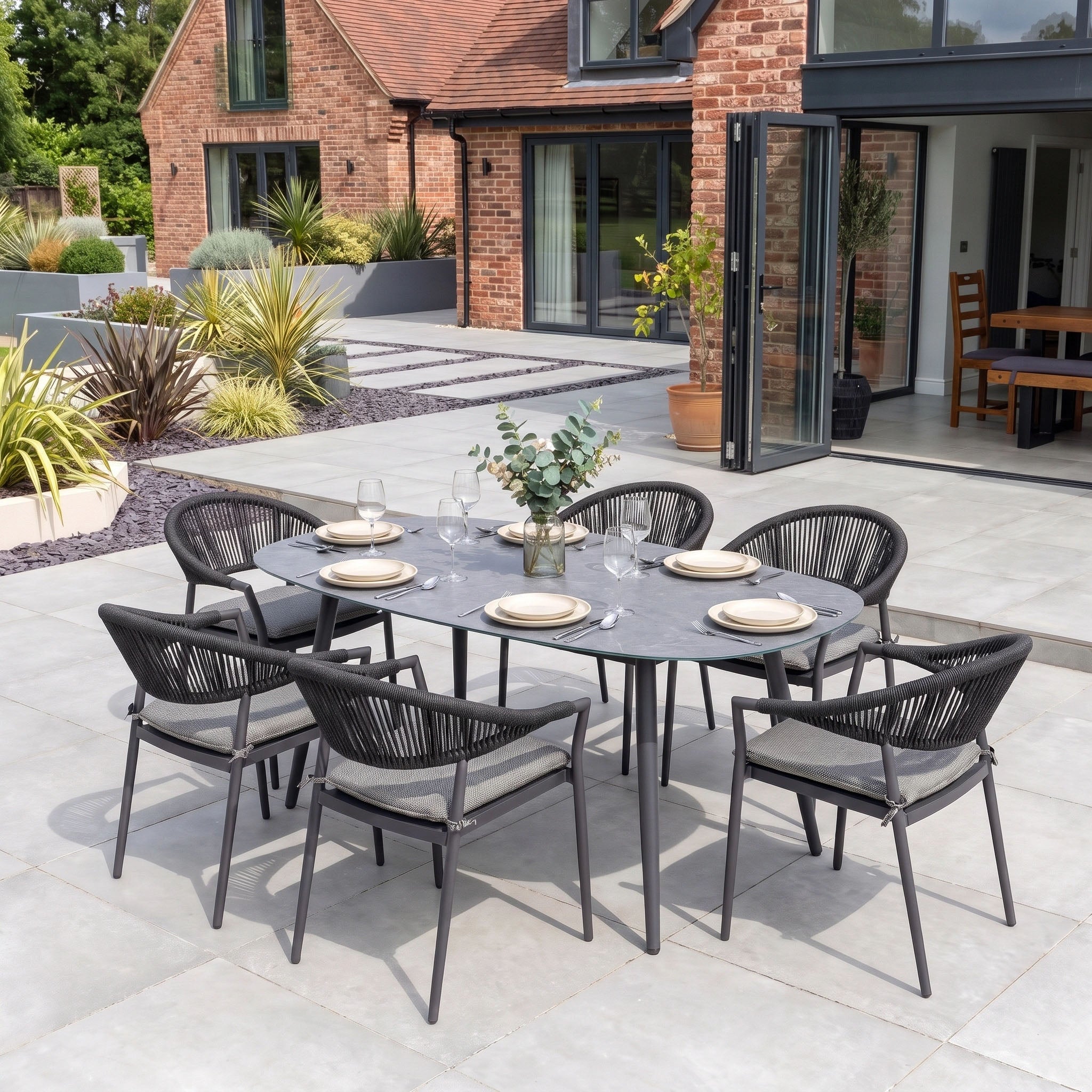 Cloverly 6 Seat Oval Dining Set with Albatross Table in Charcoal, featuring stackable dining chairs and a spacious table, perfectly arranged for outdoor dining on the patio next to a brick house.