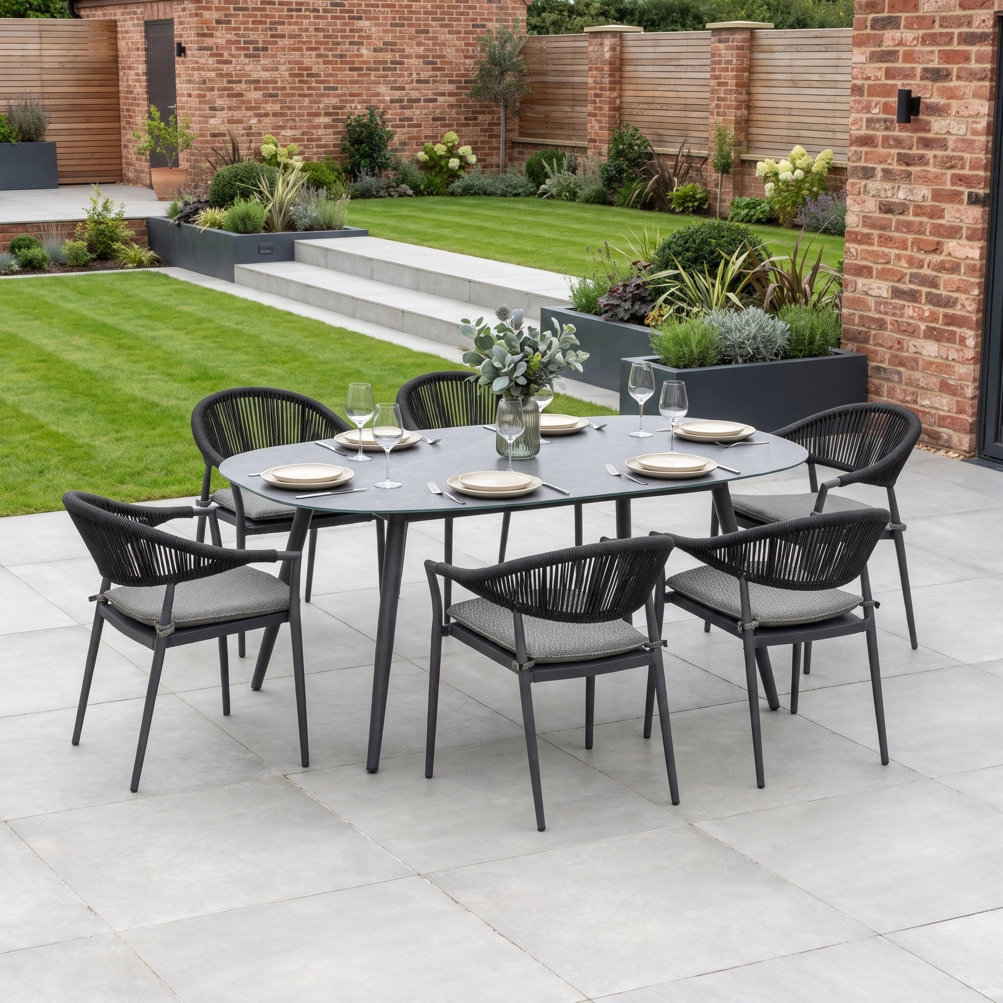 The Cloverly 6 Seat Oval Dining Set with Albatross Table in Charcoal is elegantly arranged on the patio, its stackable dining chairs overlooking a neatly landscaped garden.