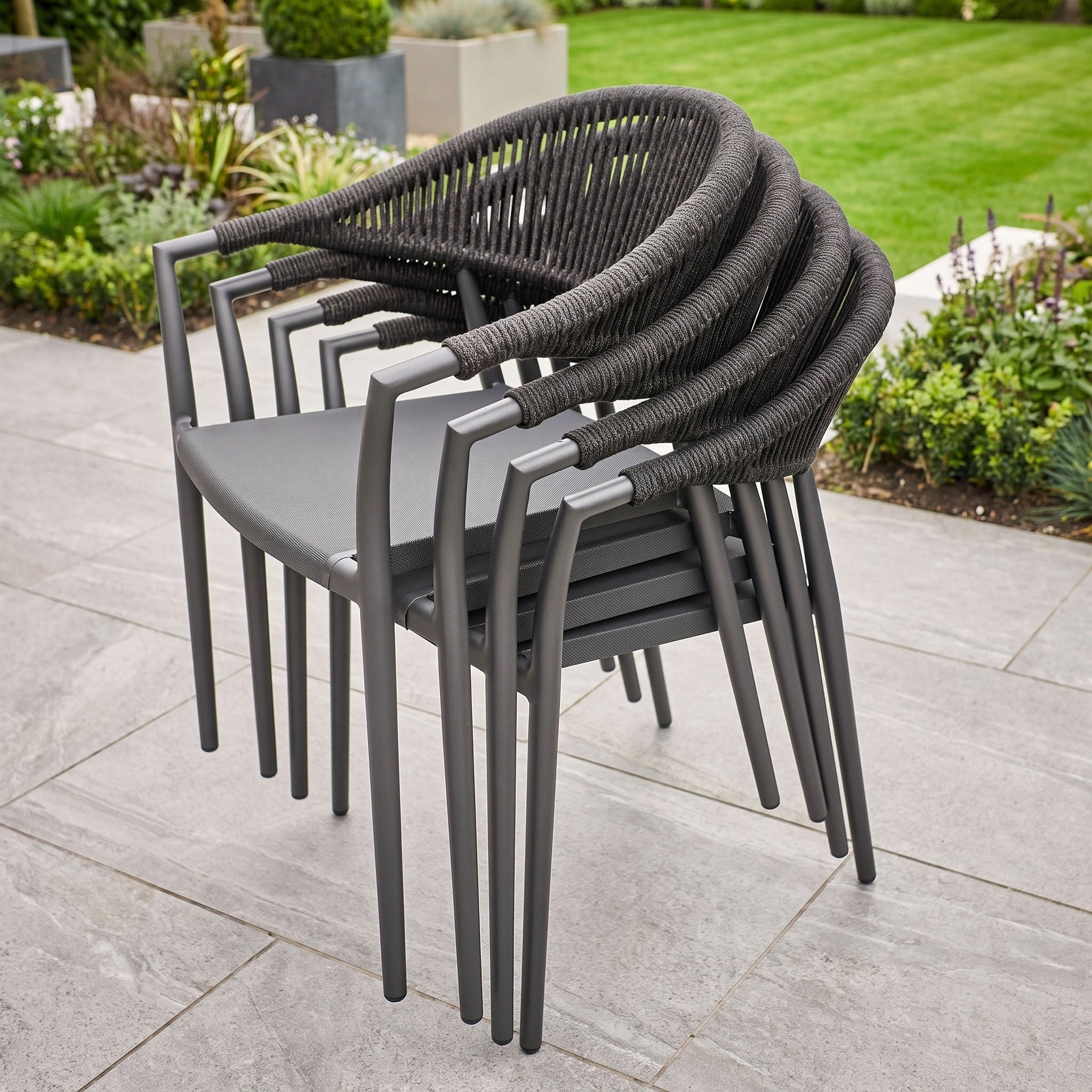 Six modern charcoal dining chairs from the Cloverly 6 Seat Oval Dining Set with Albatross Table are arranged on a tiled patio, with a garden visible in the background.