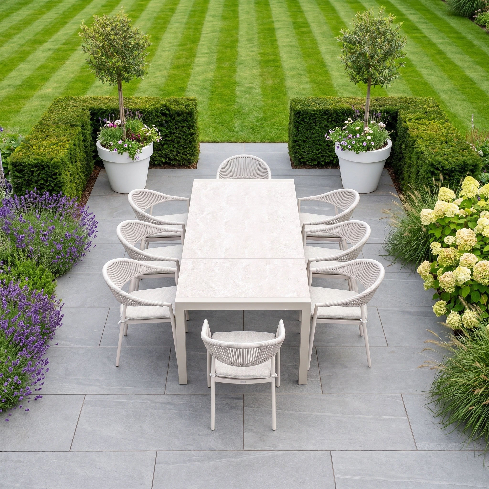 The Cloverly 8 Seat Dining Set with Ravona Extending Table in Latte, complete with eight chairs, forms an elegant outdoor arrangement surrounded by lush greenery and overlooking a neatly mowed garden lawn—perfect for patio furniture enthusiasts.