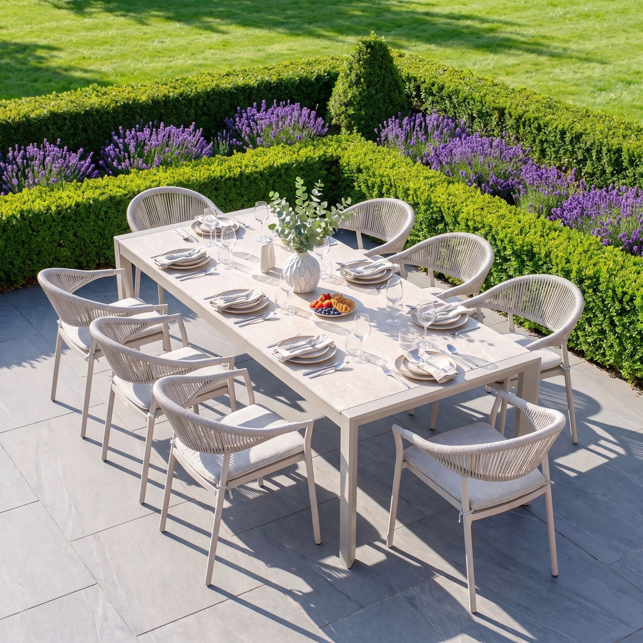 Outdoor patio furniture sets the scene for elegant gatherings with the Cloverly 8 Seat Dining Set featuring the Ravona Extending Table in Latte. The centerpiece of a spacious patio, this set is framed by lush green hedges and blooming purple flowers, providing a stylish backdrop for memorable al fresco dining experiences.