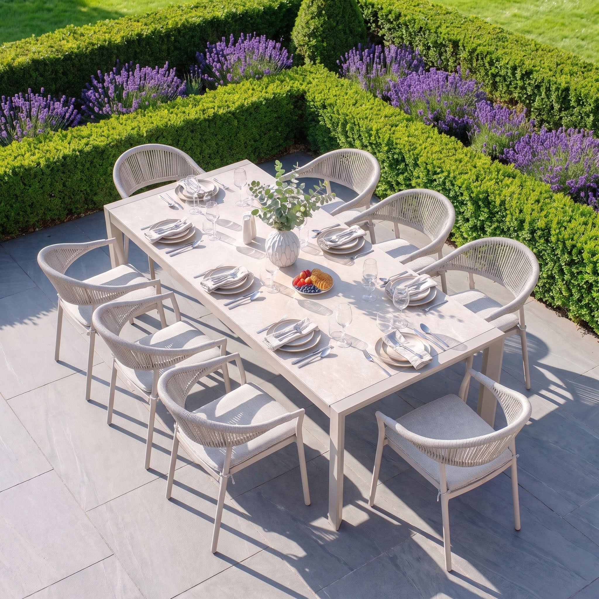 The Cloverly 8 Seat Dining Set with Ravona Extending Table in Latte is featured outdoors, surrounded by green hedges and blooming purple flowers.