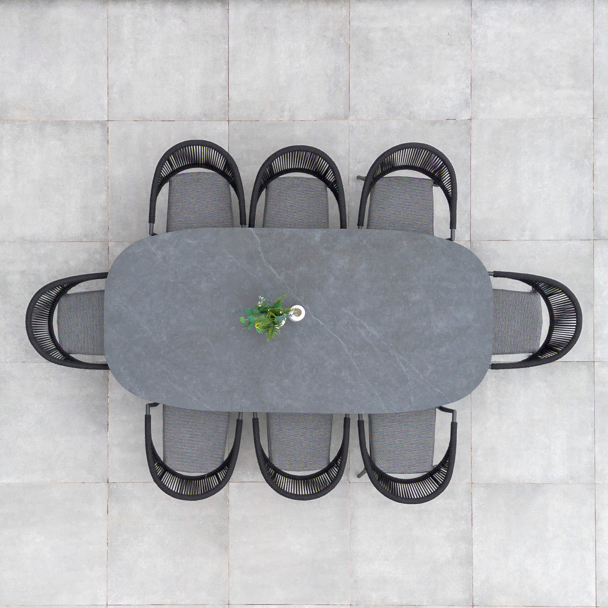 Top view of the Cloverly 8 Seat Rope Oval Dining Set with Ceramic Table in Charcoal outdoors, featuring a maintenance-free aluminum frame and a ceramic glass table, surrounded by six black chairs with a small potted plant in the center.