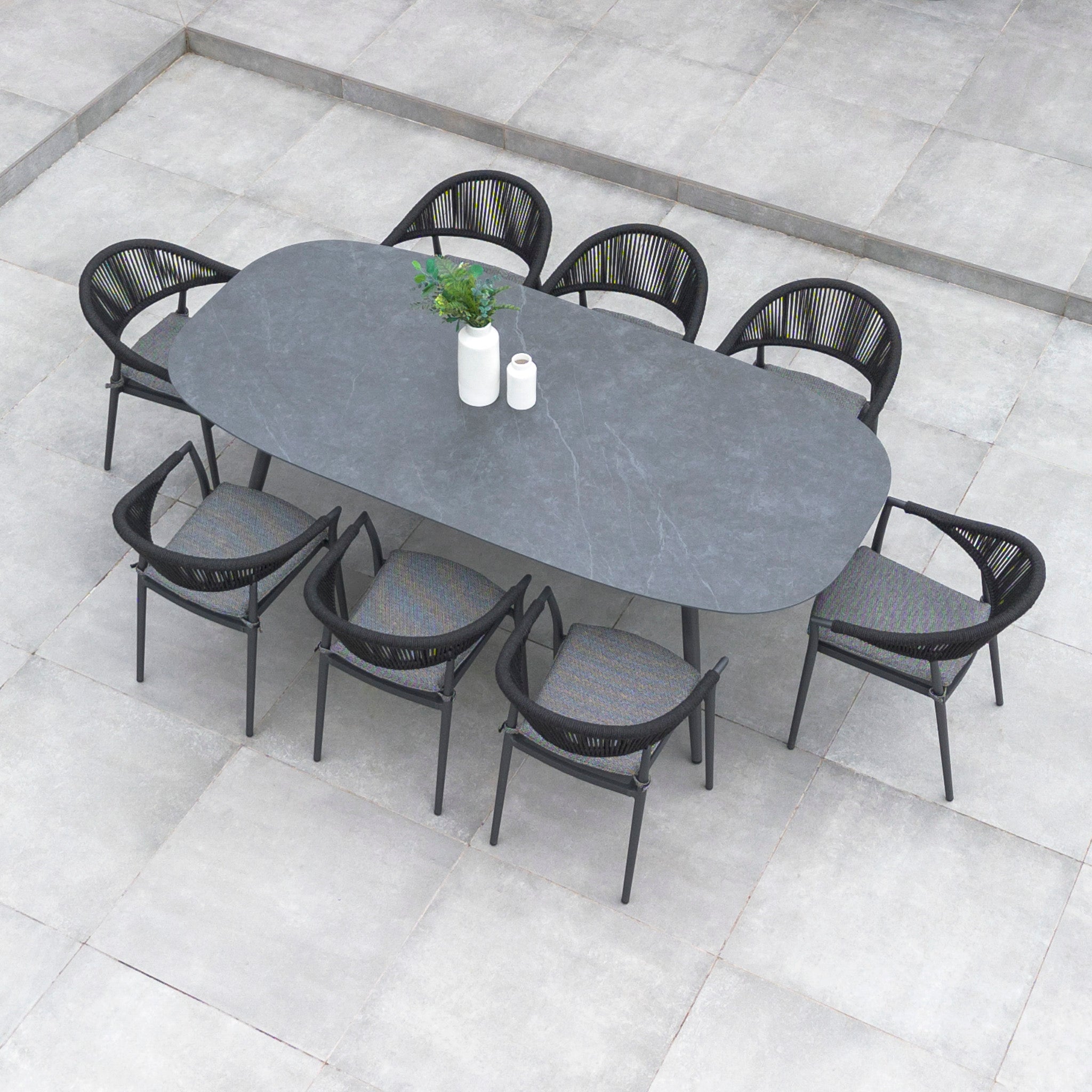 Top view of a Cloverly 8 Seat Rope Oval Dining Set with Ceramic Table in Charcoal, placed on a tiled floor, featuring a plant and two bottles on the maintenance-free aluminium table with ceramic glass.