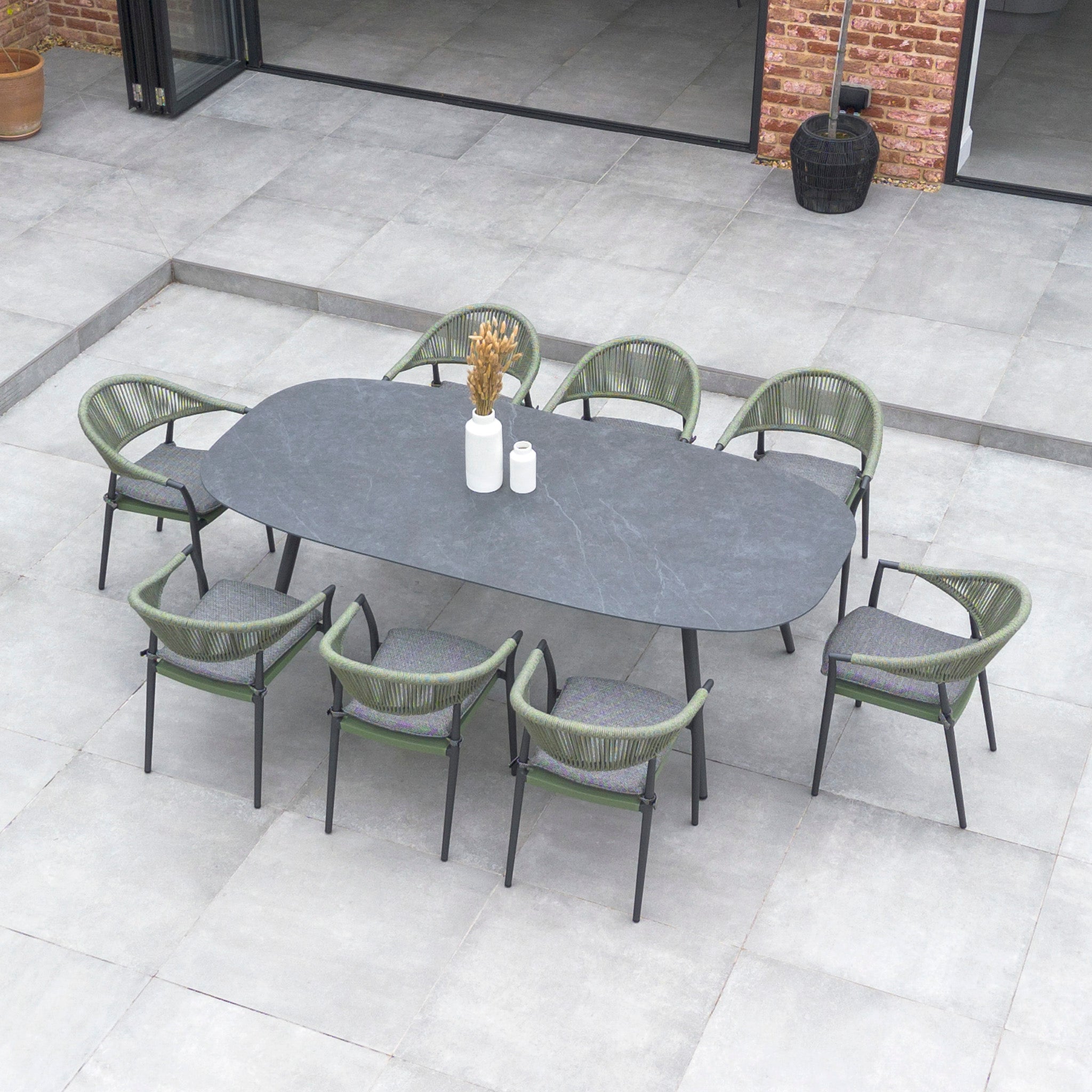 A Cloverly 8 Seat Rope Oval Dining Set with Ceramic Table in Olive Green, featuring stackable woven chairs, surrounds the table on a tiled patio outside a brick-walled building.