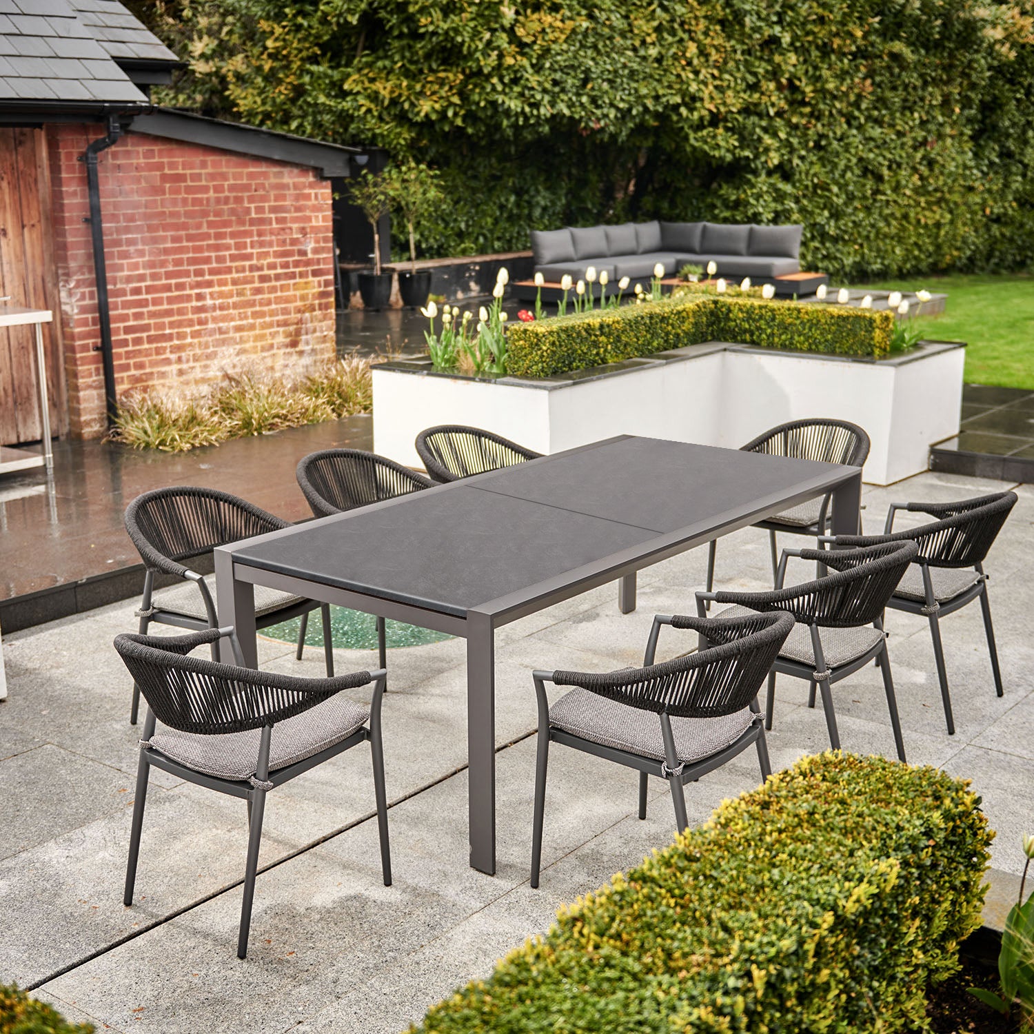 Modern outdoor patio with a Cloverly 8 Seat Rectangular Extending Dining Set with Ceramic Table in Charcoal featuring weather-proof aluminium, eight chairs with removable sun-proof cushions, and lush greenery in the background.